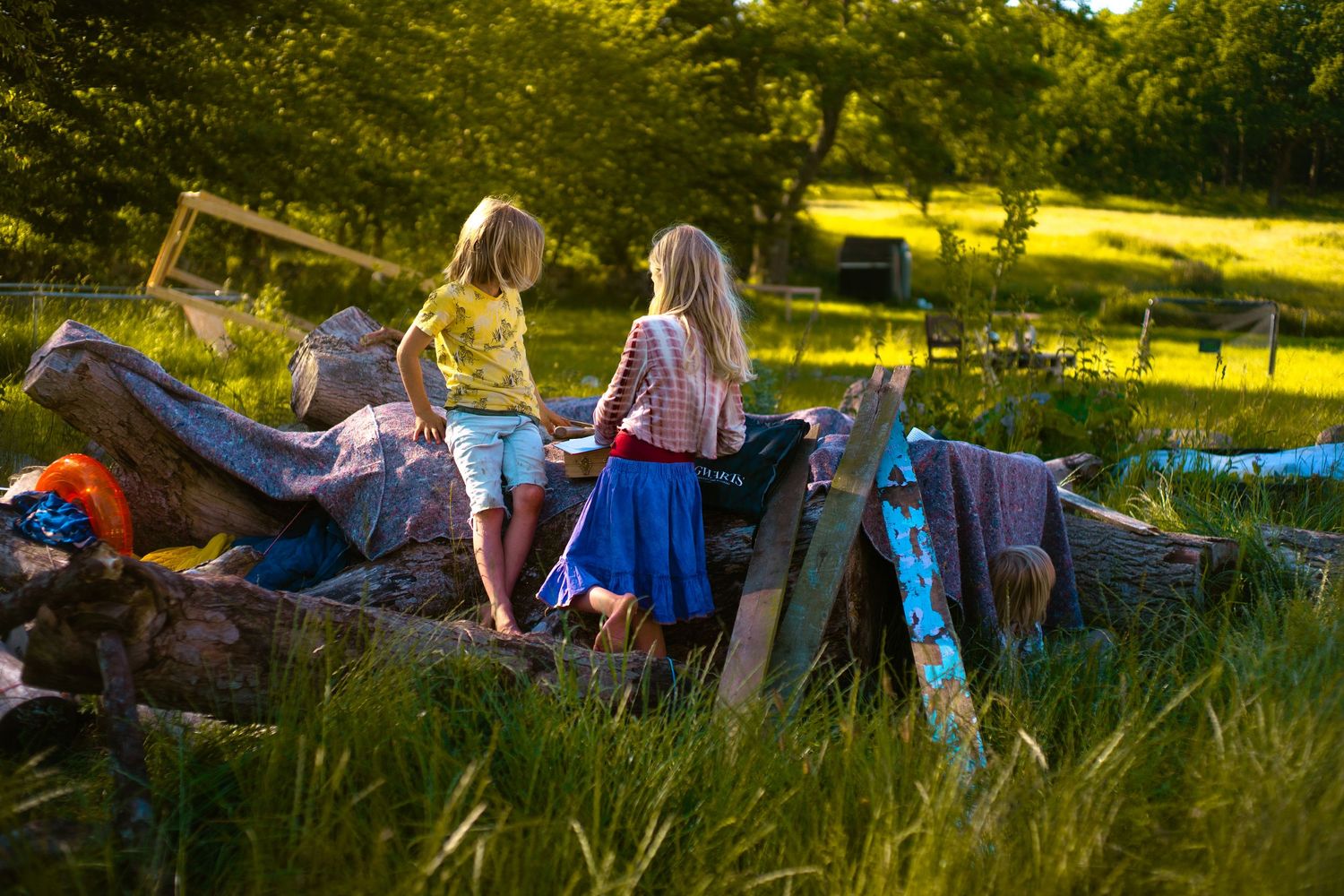 Two children explore a rustic outdoor pile of wood and debris in a lush green meadow during golden hour.
