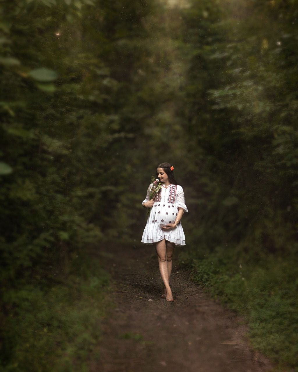 A figure in a white dress walks barefoot along a wooded dirt path on a misty day.