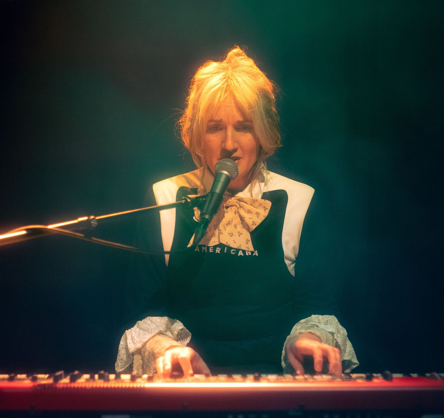 Blonde musician playing red keyboard and singing into microphone on dimly lit stage wearing black and white outfit.
