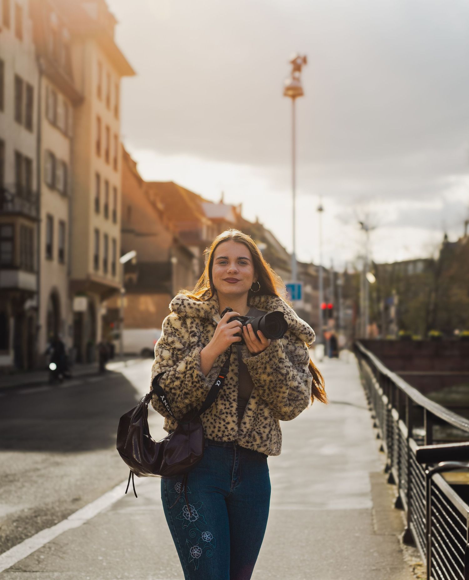 Young woman in leopard print jacket holding a camera walks along a European city street at golden hour.