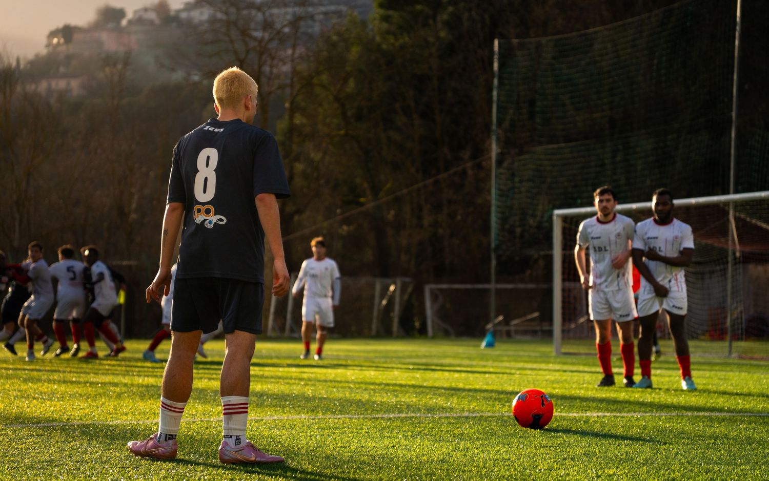 Player wearing number 8 jersey prepares to kick a red soccer ball during a sunny outdoor match on a green field.