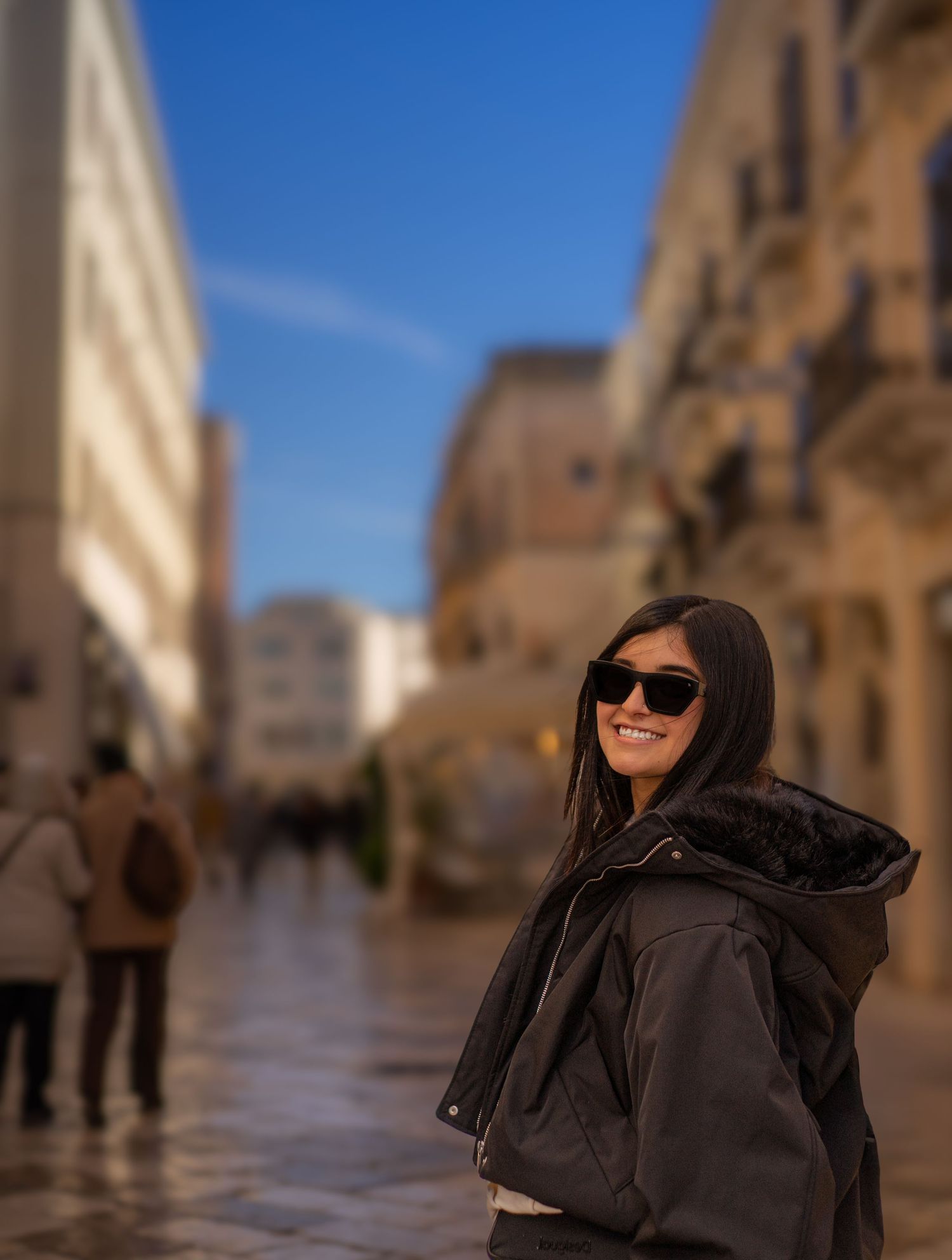 Smiling young woman in black jacket and sunglasses posing on a sunny European city street.