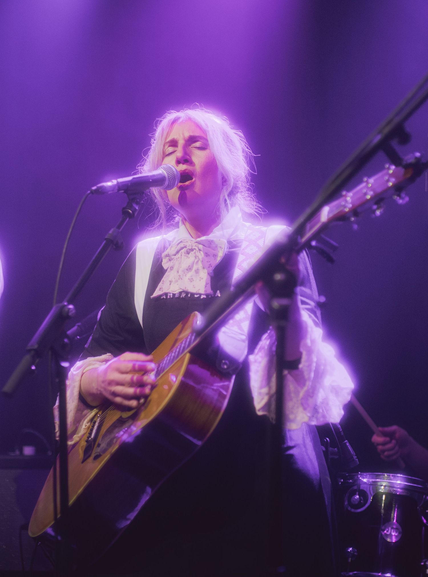 Blonde female vocalist playing acoustic guitar on stage under purple lights at a live concert performance.