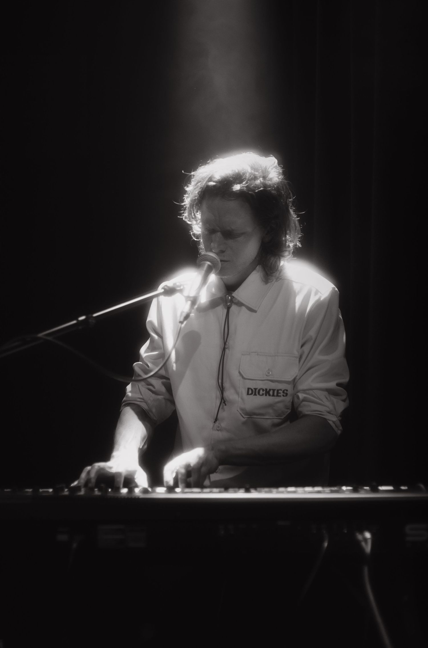 Black and white photo of a male musician playing keyboard and singing into a microphone on a dimly lit stage.