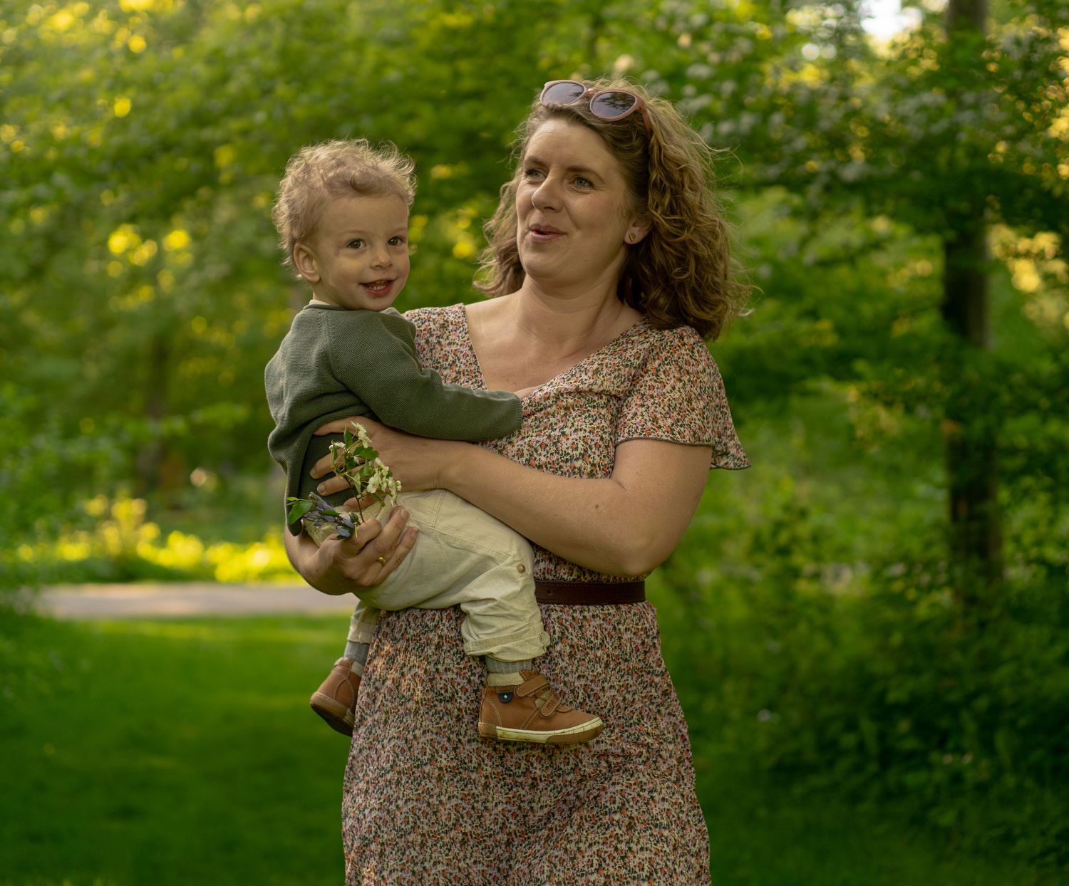 A woman in a floral dress holds a toddler boy outdoors in a lush green park setting.