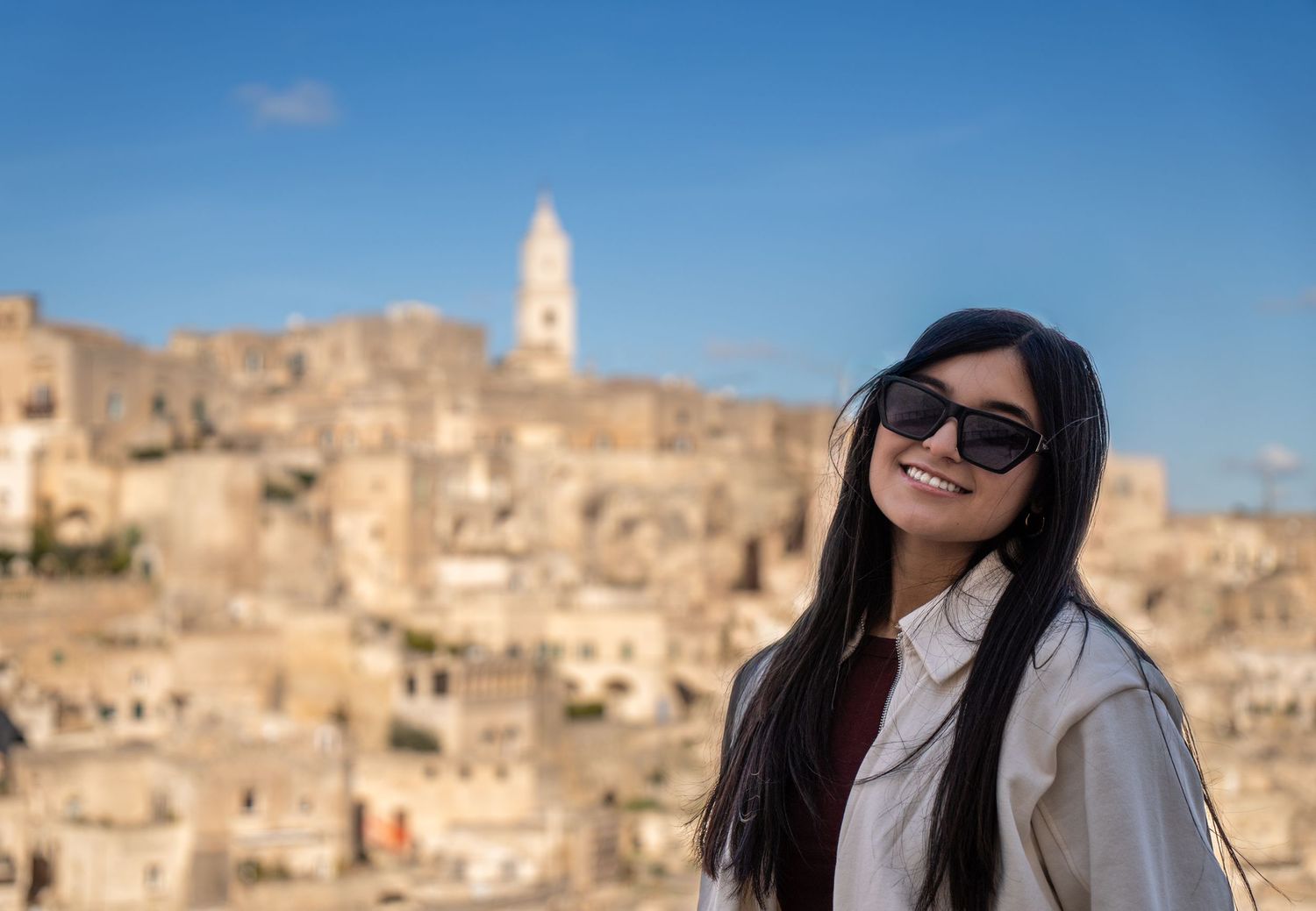 Young woman with sunglasses smiling in front of the ancient cave city of Matera, Italy on a sunny day.
