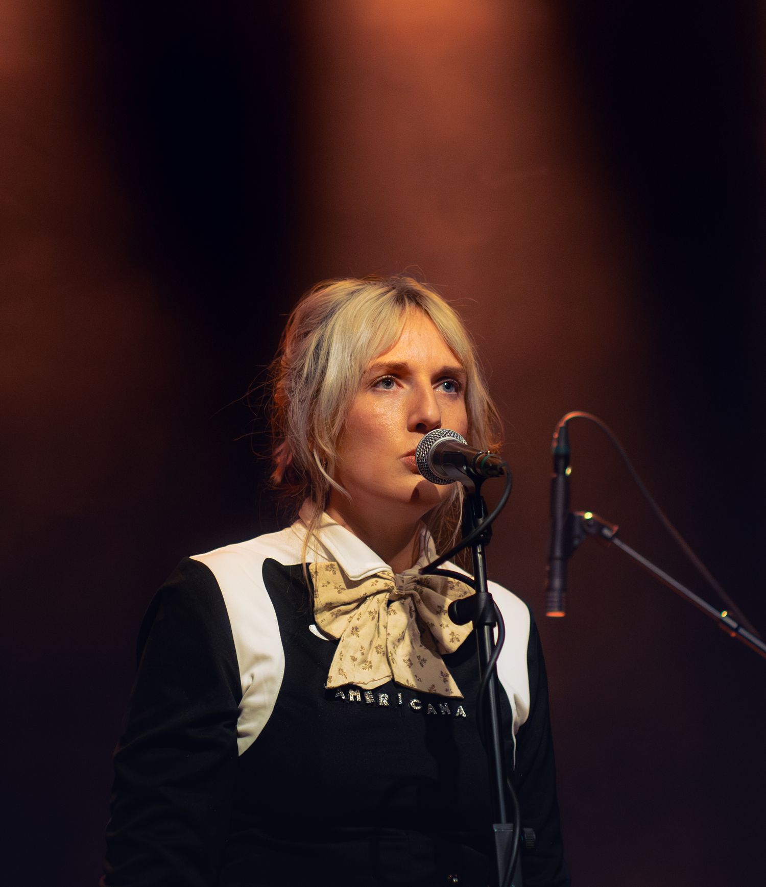 Blonde female singer performing on stage at a concert, wearing a black jacket with a scarf near a microphone.