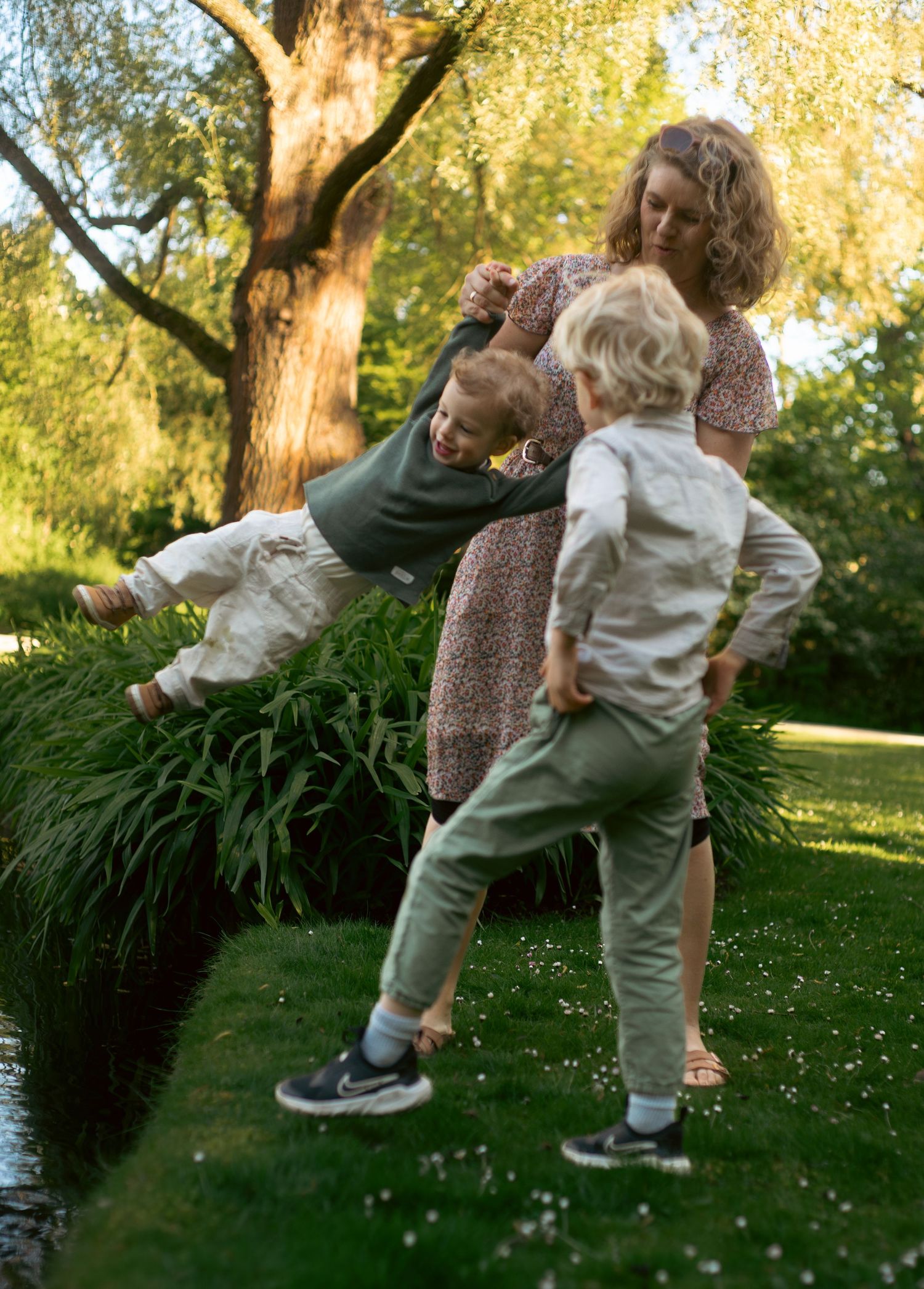 A mother playfully swings her young son in a lush green garden while another child watches nearby.