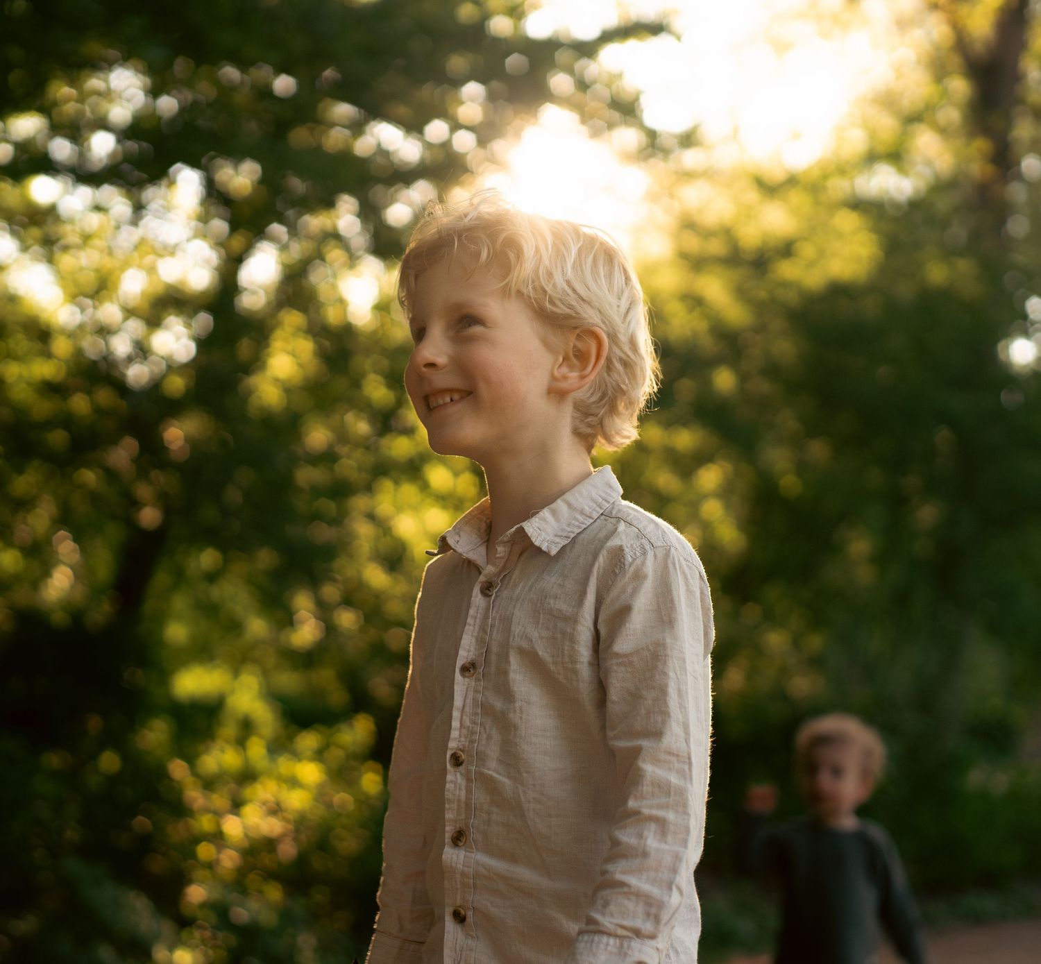 A smiling blonde boy in a linen shirt stands outdoors in golden sunlight with lush green trees in the background.