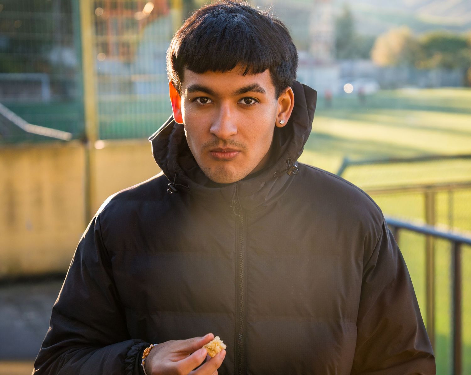 Young man in black puffer jacket holding food near a sports field in warm sunlight.