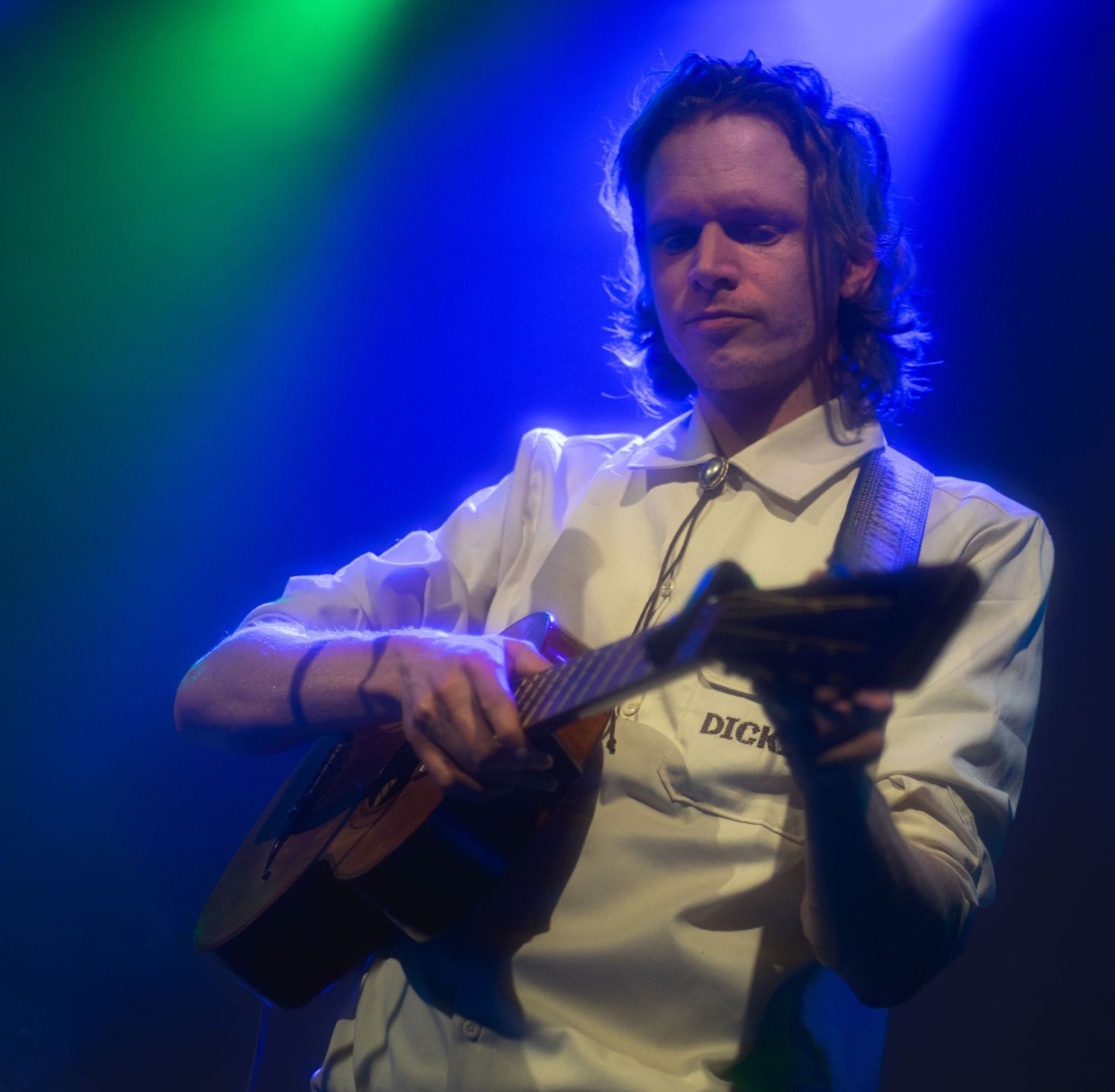 Musician playing acoustic guitar on stage under colorful green and blue spotlights during a live performance.