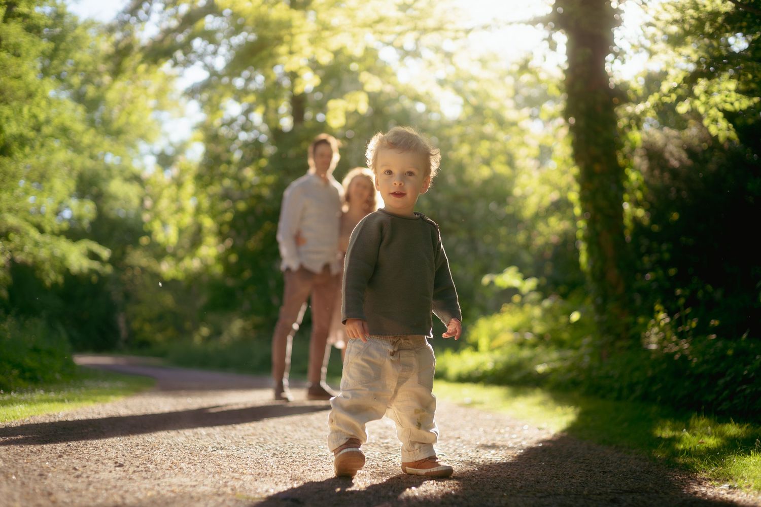 Toddler running on a sunny park path with parents walking in the background among lush green trees.