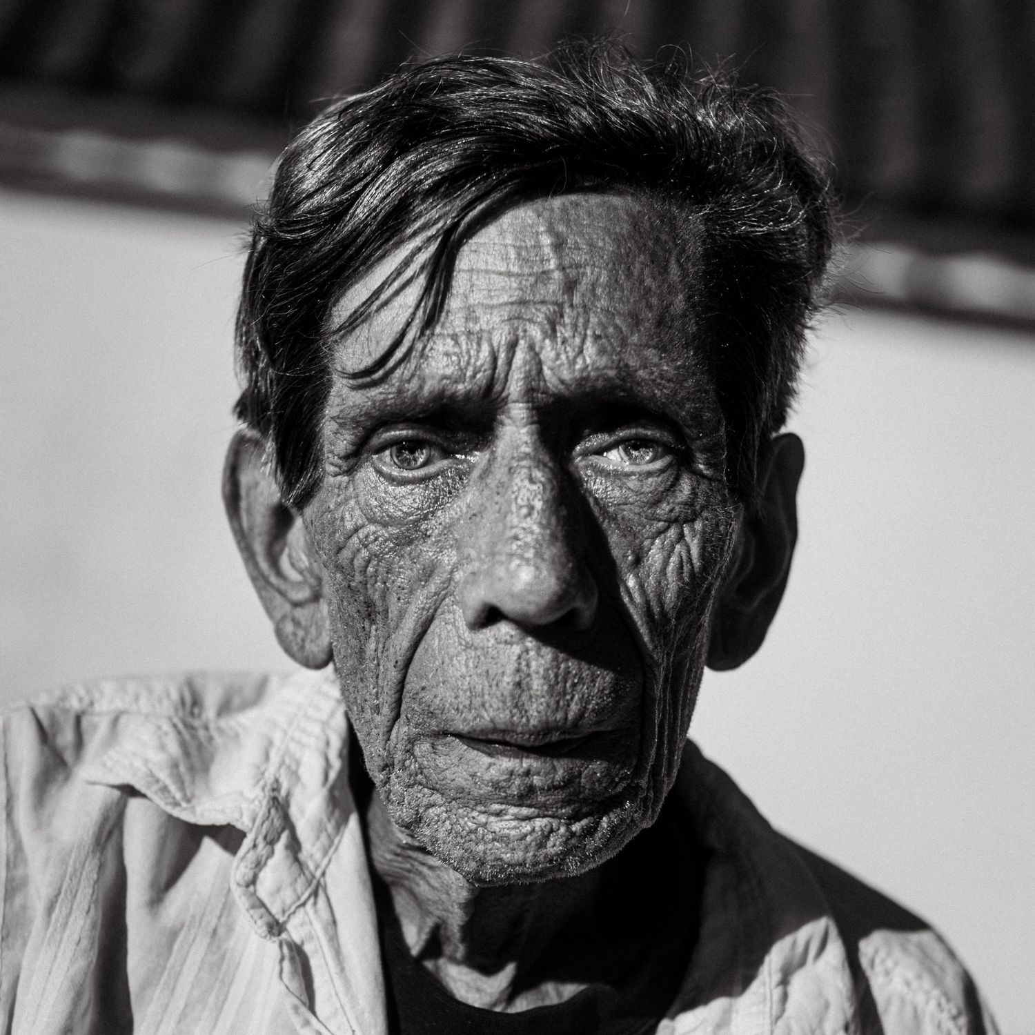 Black and white portrait of an elderly man with deeply wrinkled skin, wearing a light shirt, looking directly at camera.