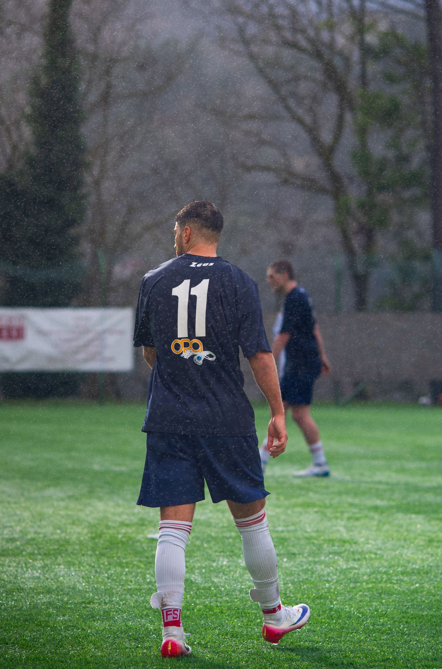 Soccer player wearing number 11 navy jersey and white socks stands on green field during a rainy match.