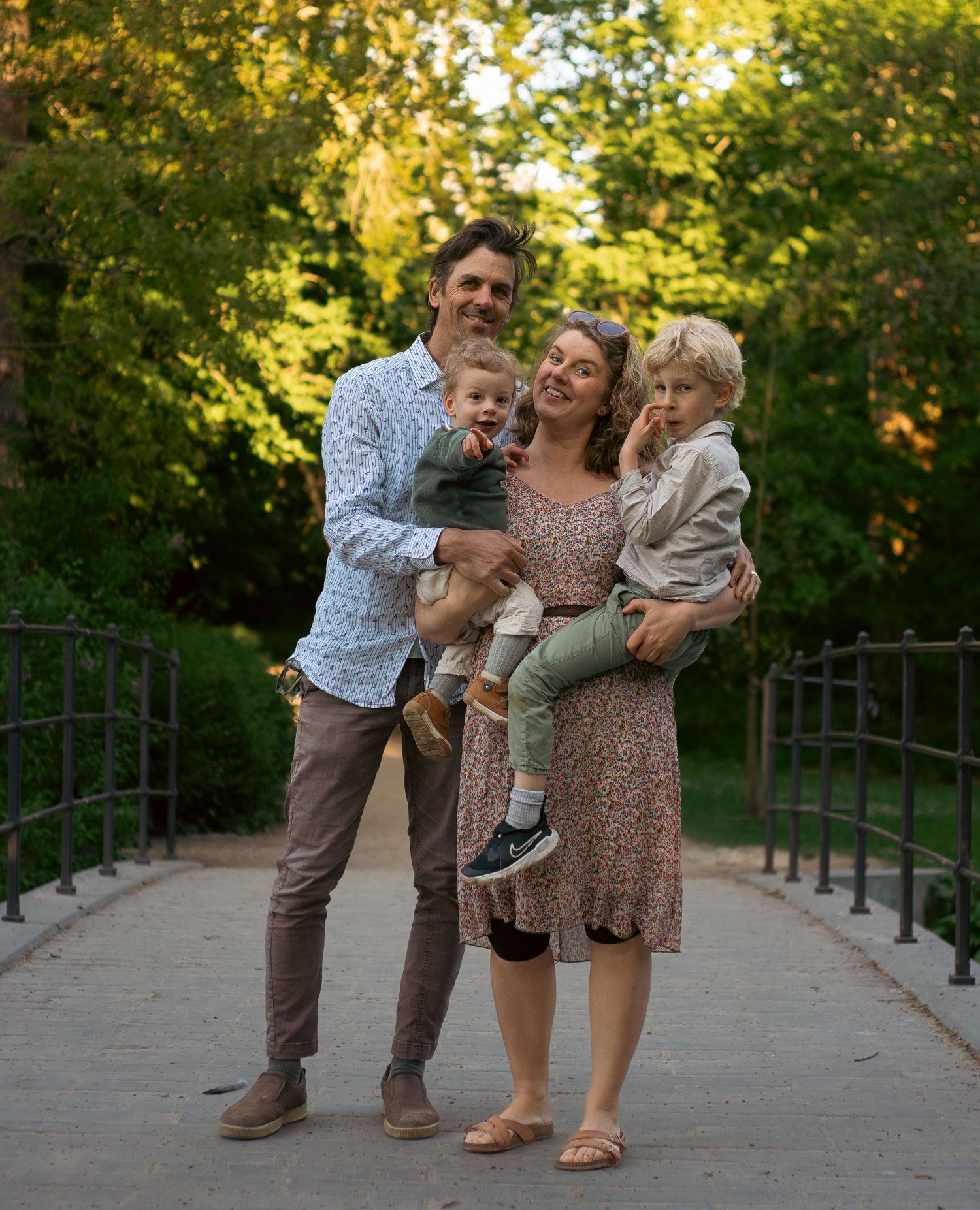A happy family of four poses on a park bridge, parents holding two young children, surrounded by lush green trees.
