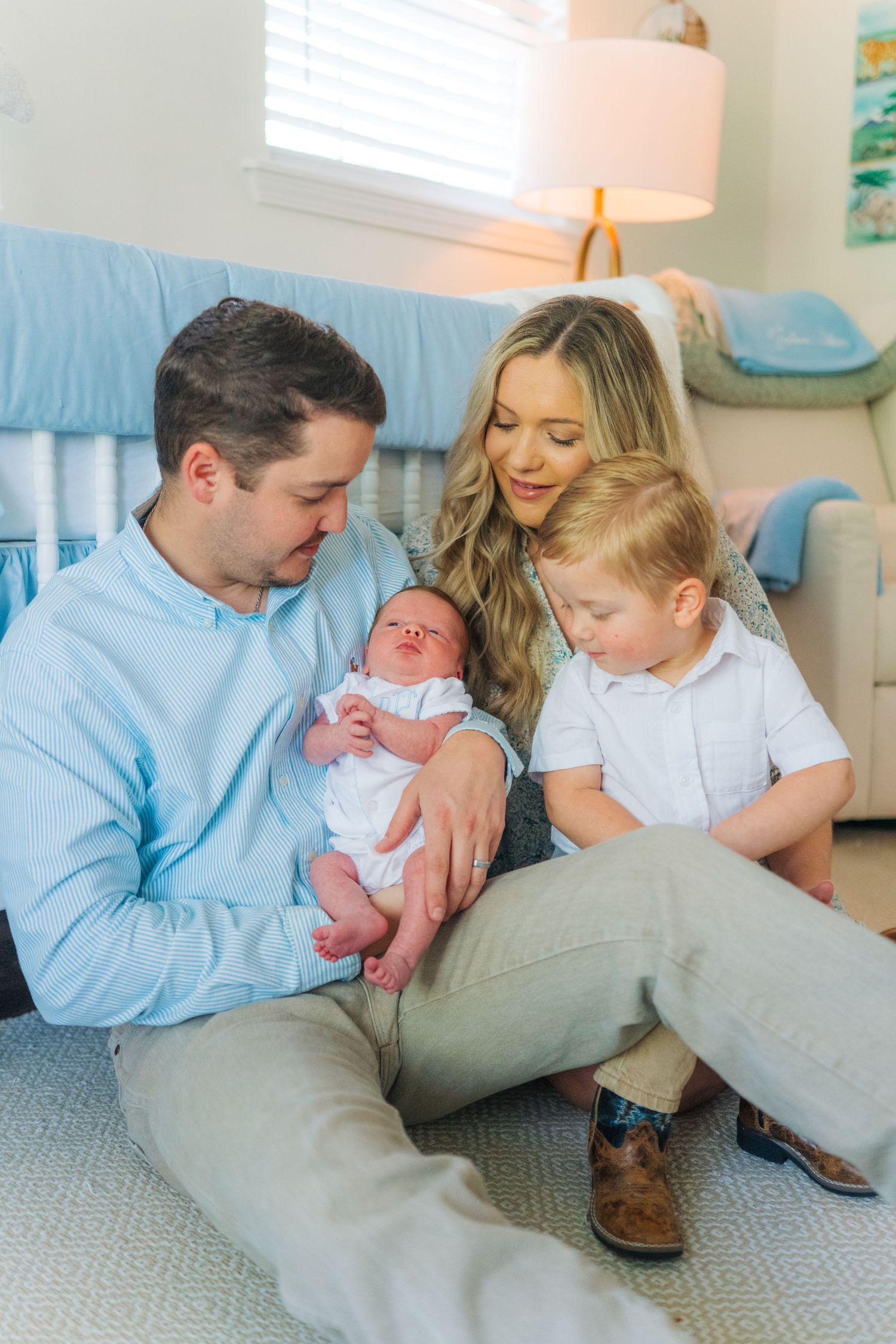 A young family sits on a nursery floor, parents gazing lovingly at their newborn baby while toddler son looks on.
