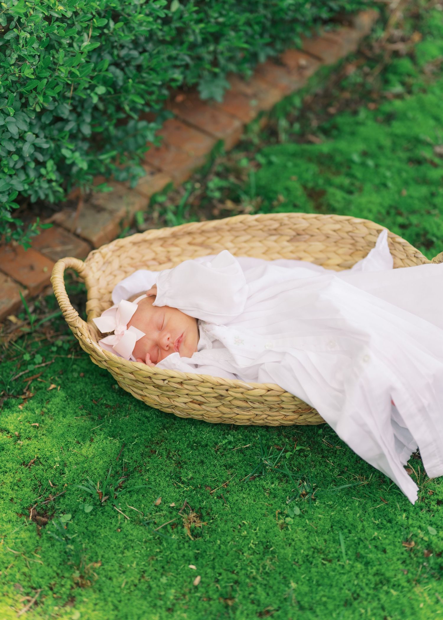 Sleeping newborn baby wrapped in white swaddle lying in a wicker basket on green grass outdoors.