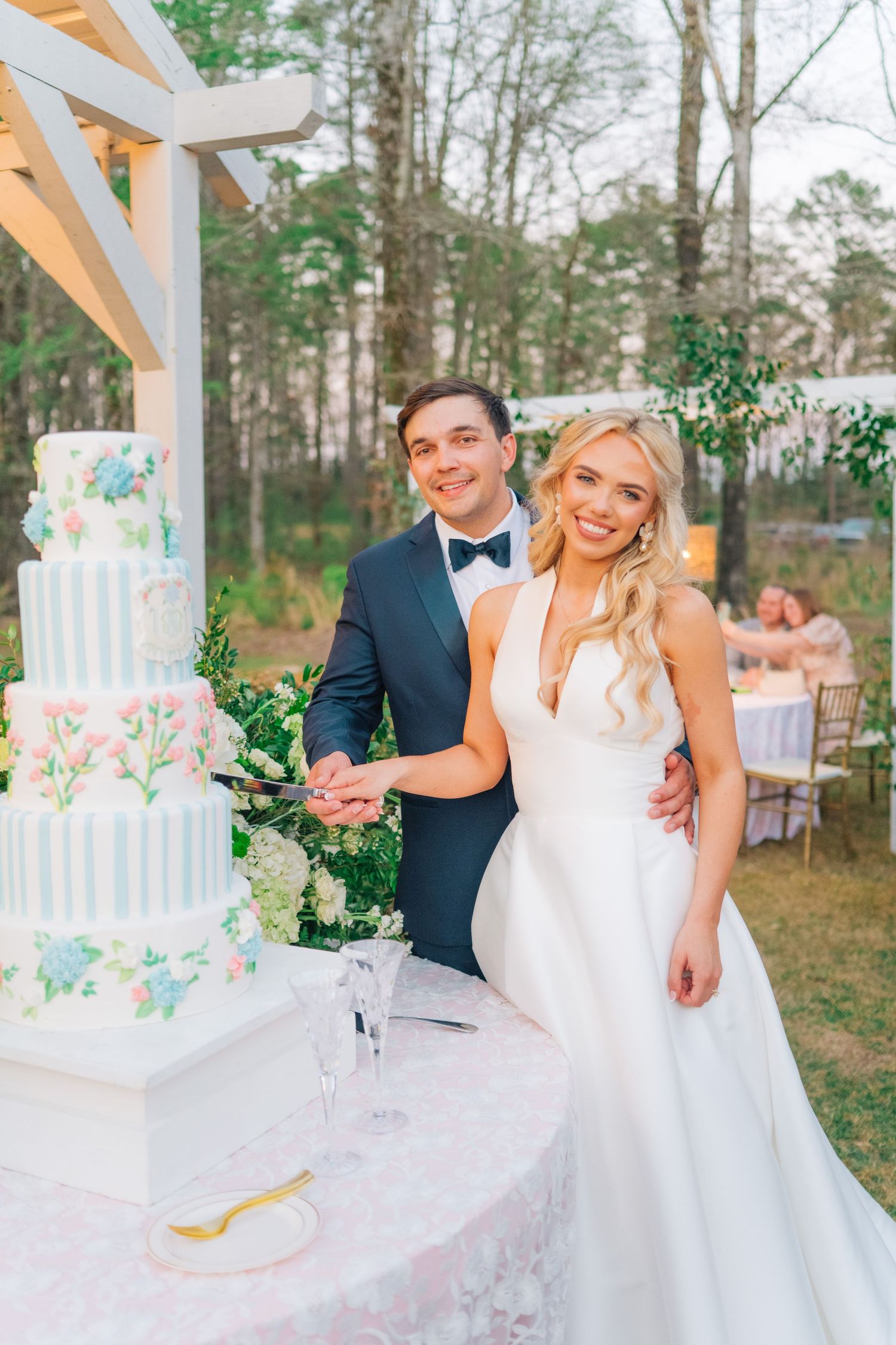 Bride and groom smiling while cutting their elegant four-tier floral wedding cake outdoors at a garden reception.