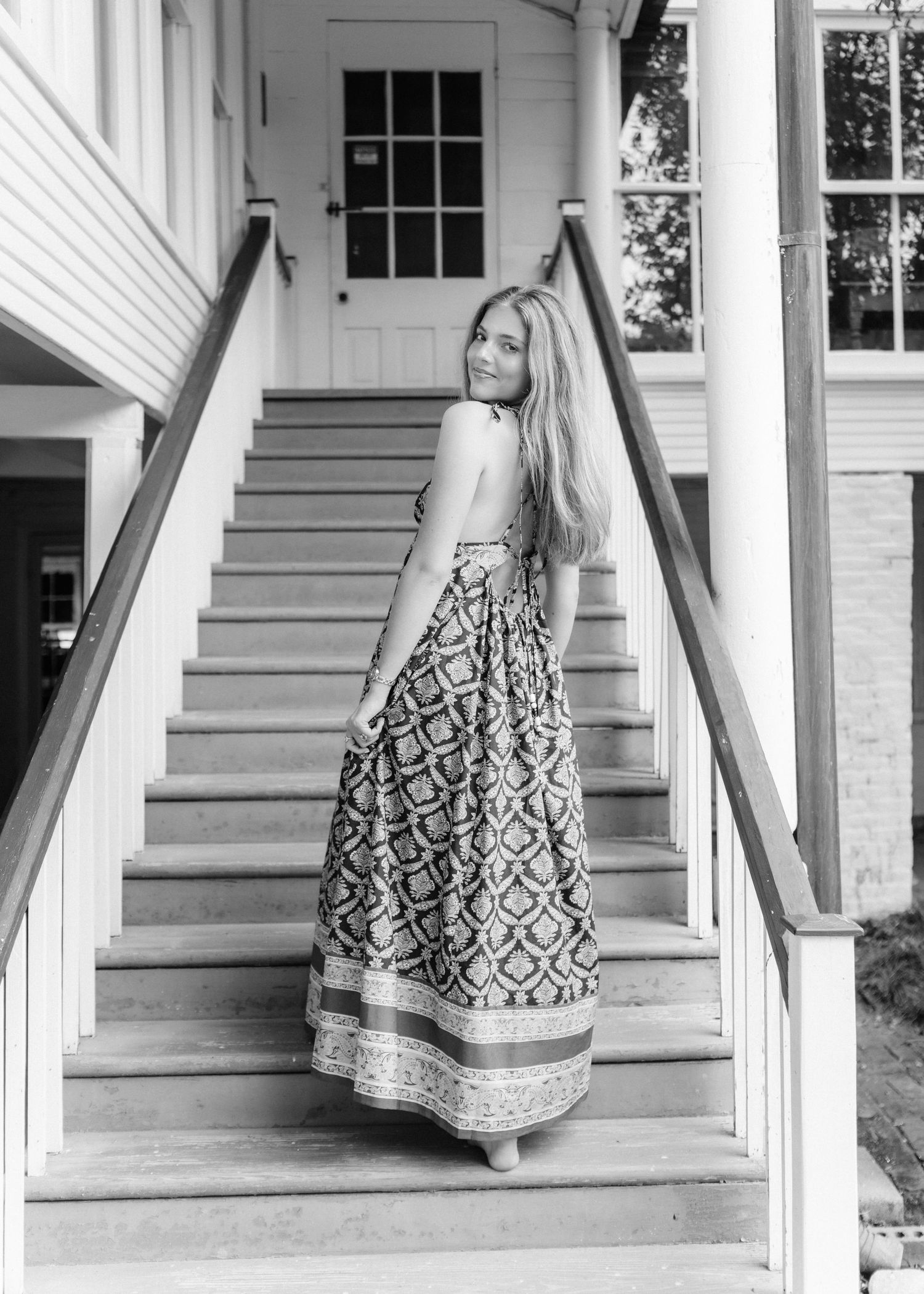 Young woman in backless patterned maxi dress looks back while standing on white porch staircase.