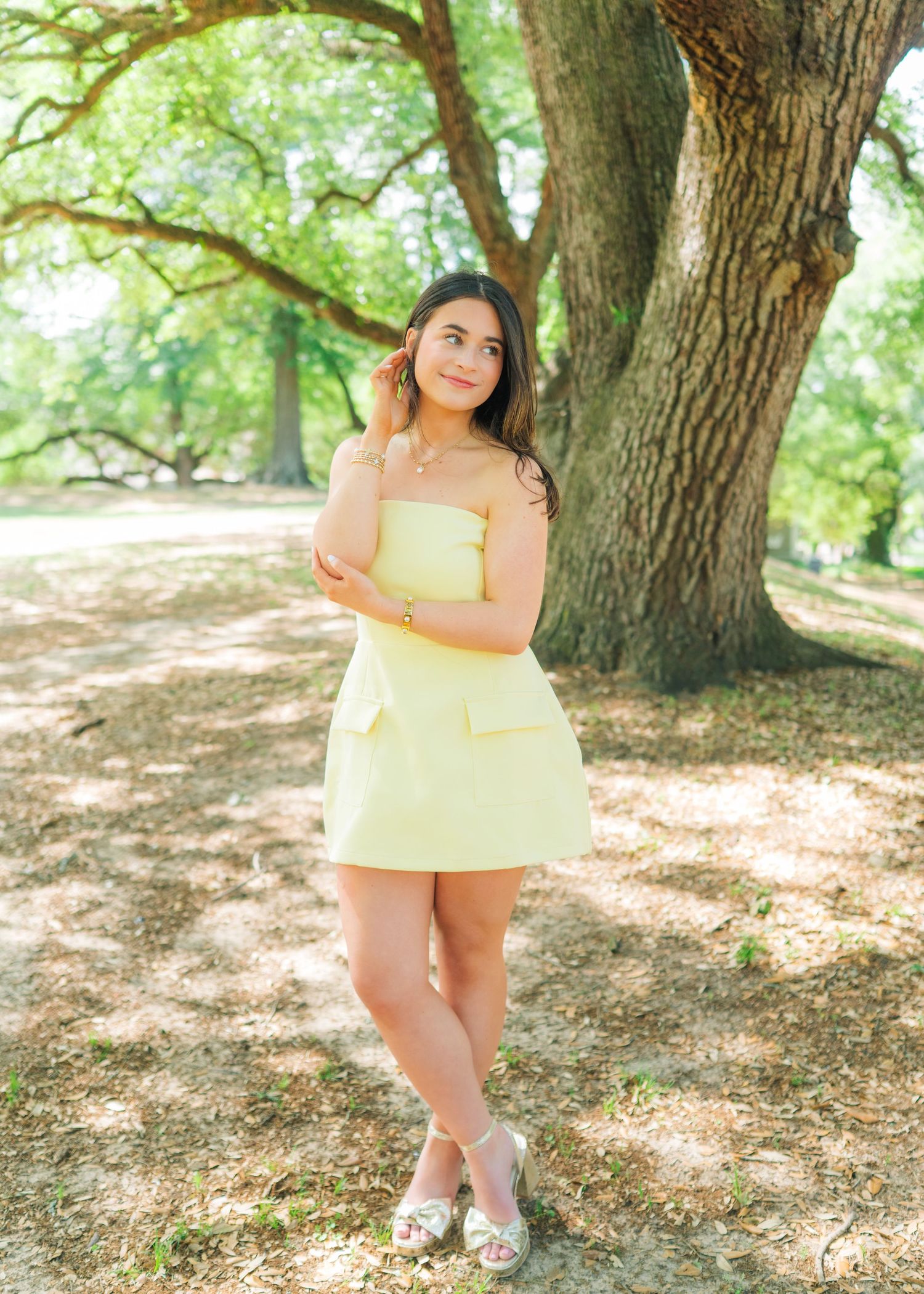 Young woman in yellow strapless dress leaning against large oak tree in sunny park setting.
