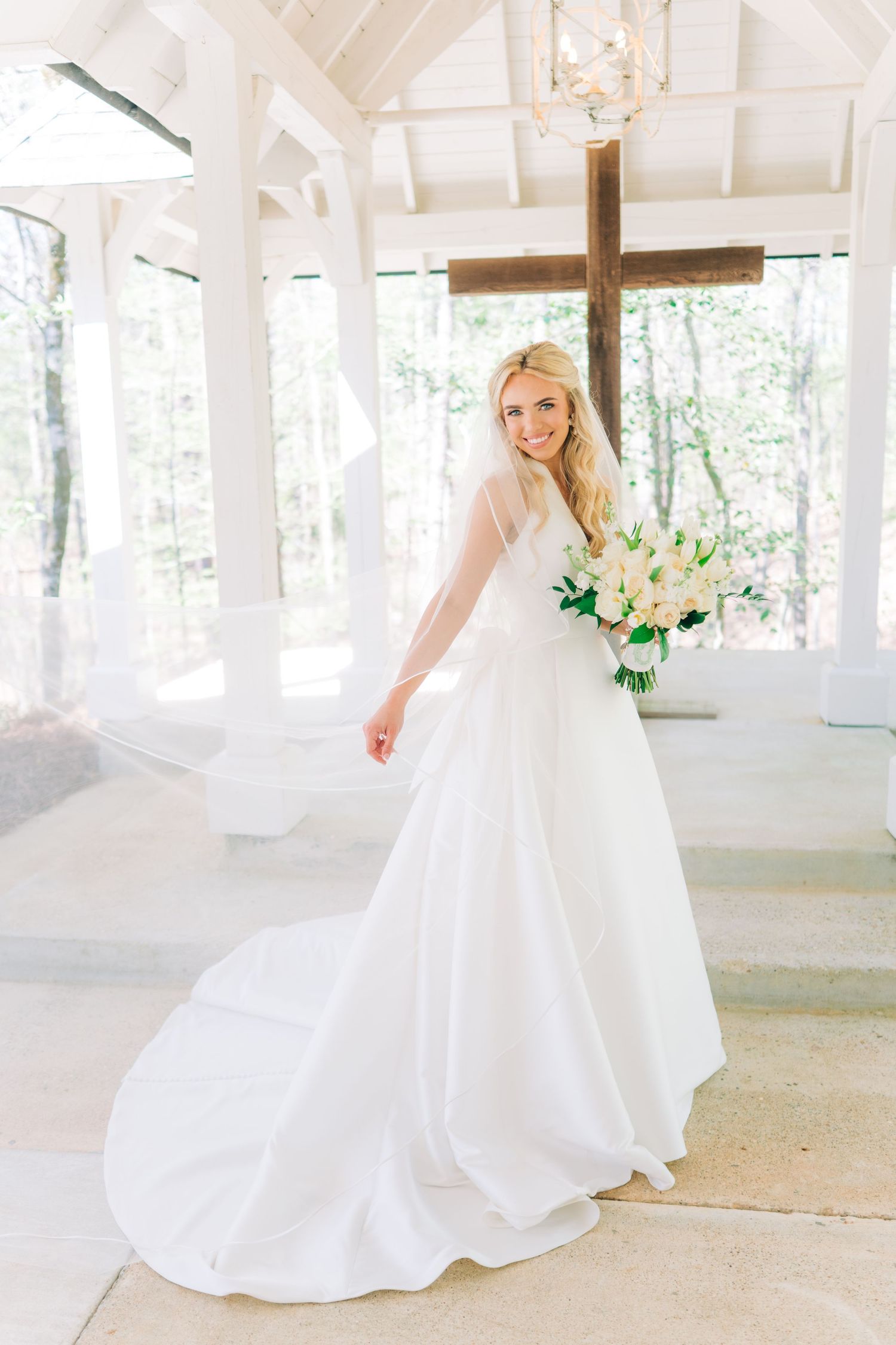 Bride in elegant white ball gown holding floral bouquet stands before wooden cross in bright white chapel.