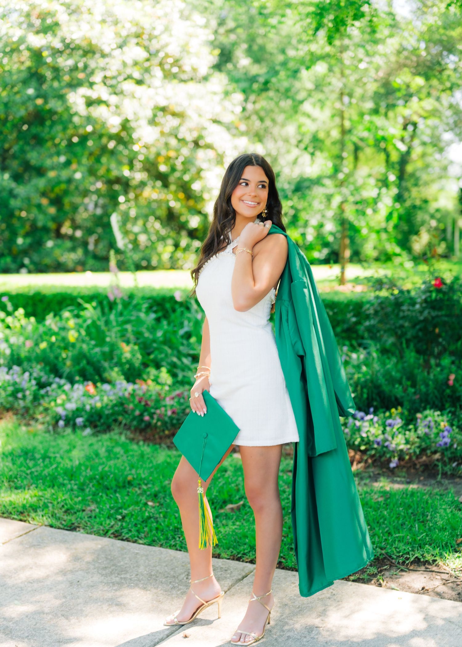 Graduate in white dress holding green cap and gown in sunny outdoor garden.