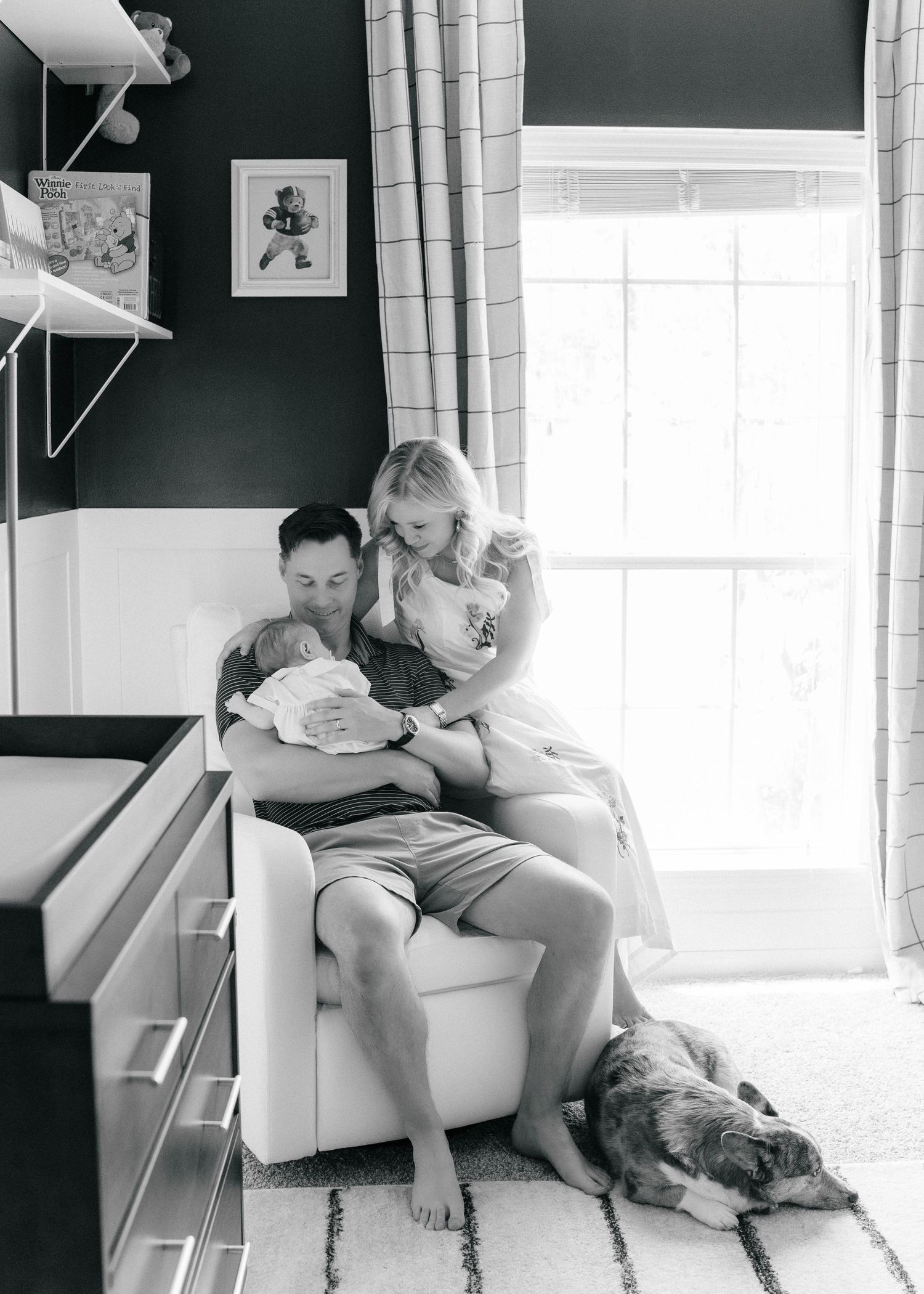Black and white photo of a couple sitting in a chair with their newborn baby and dog resting on the floor beside them.