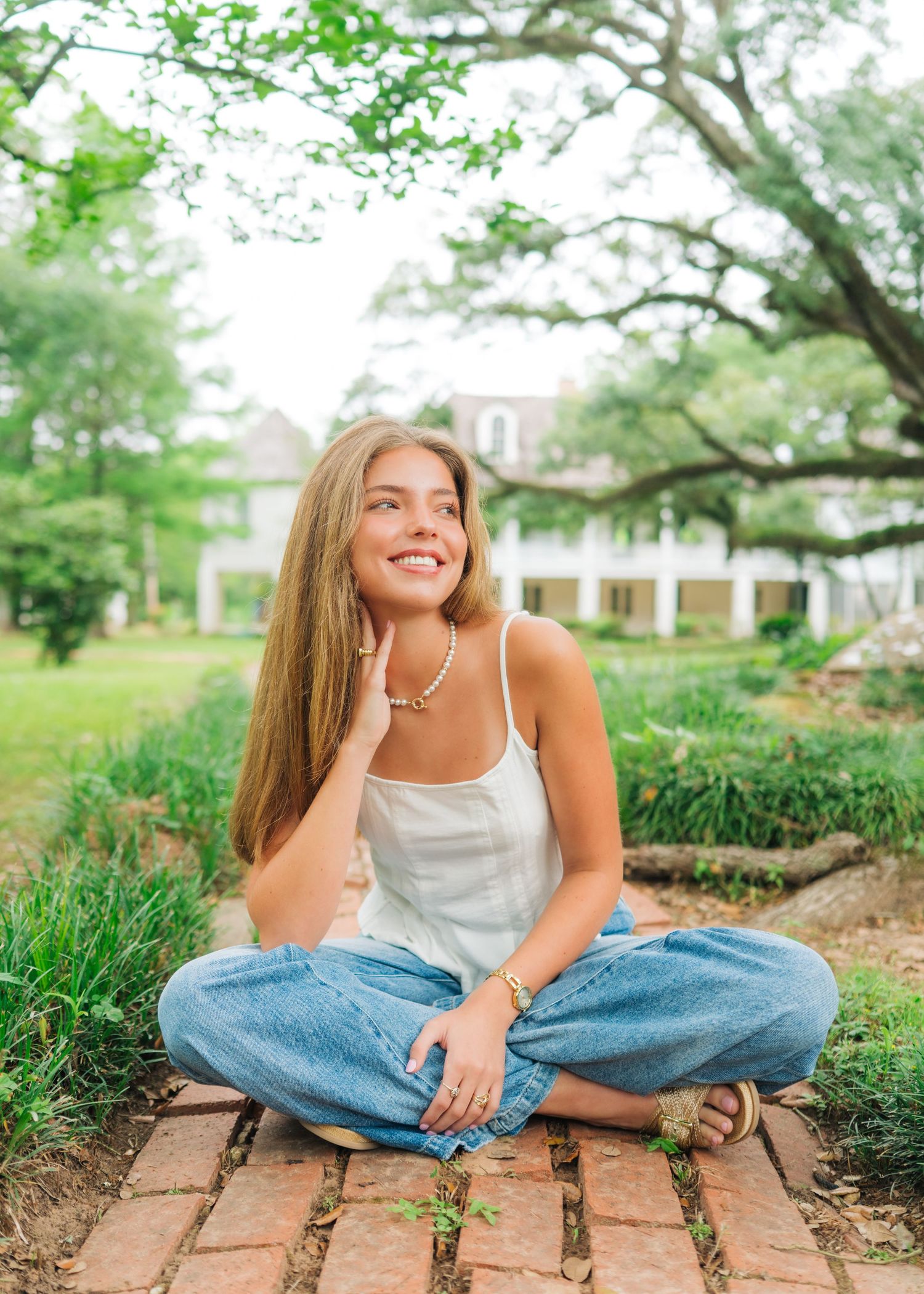 Young woman in white top and jeans sitting cross-legged on brick path, smiling outdoors near lush greenery.