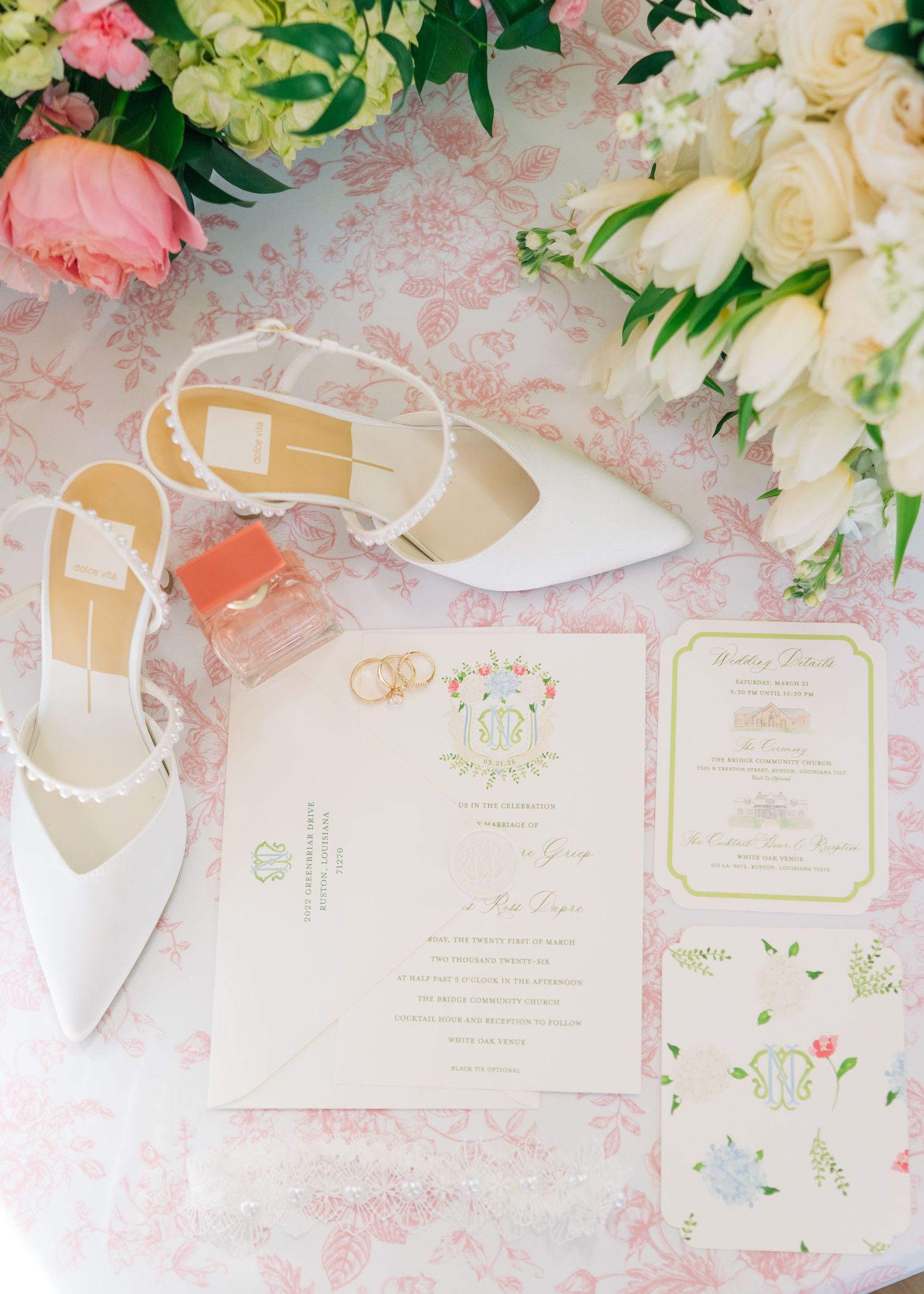 Elegant wedding flat lay with white pearl heels, gold rings, floral invitations, and pink roses on a toile background.