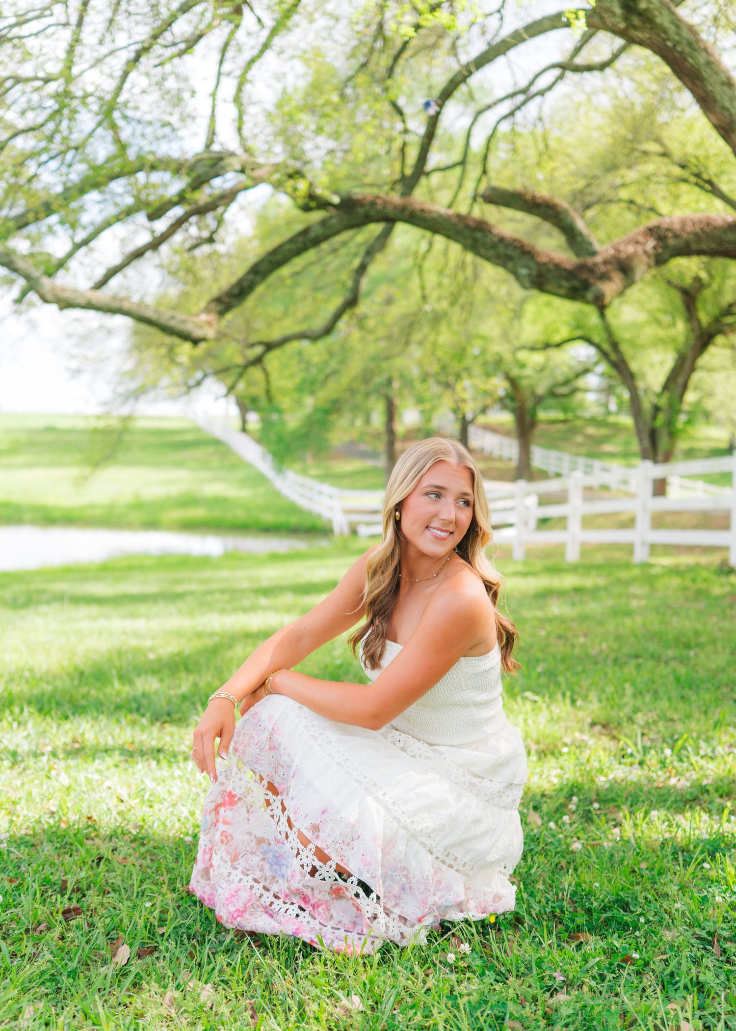 Blonde teen in white floral maxi dress sits on green grass beneath sprawling oak tree for senior photos.
