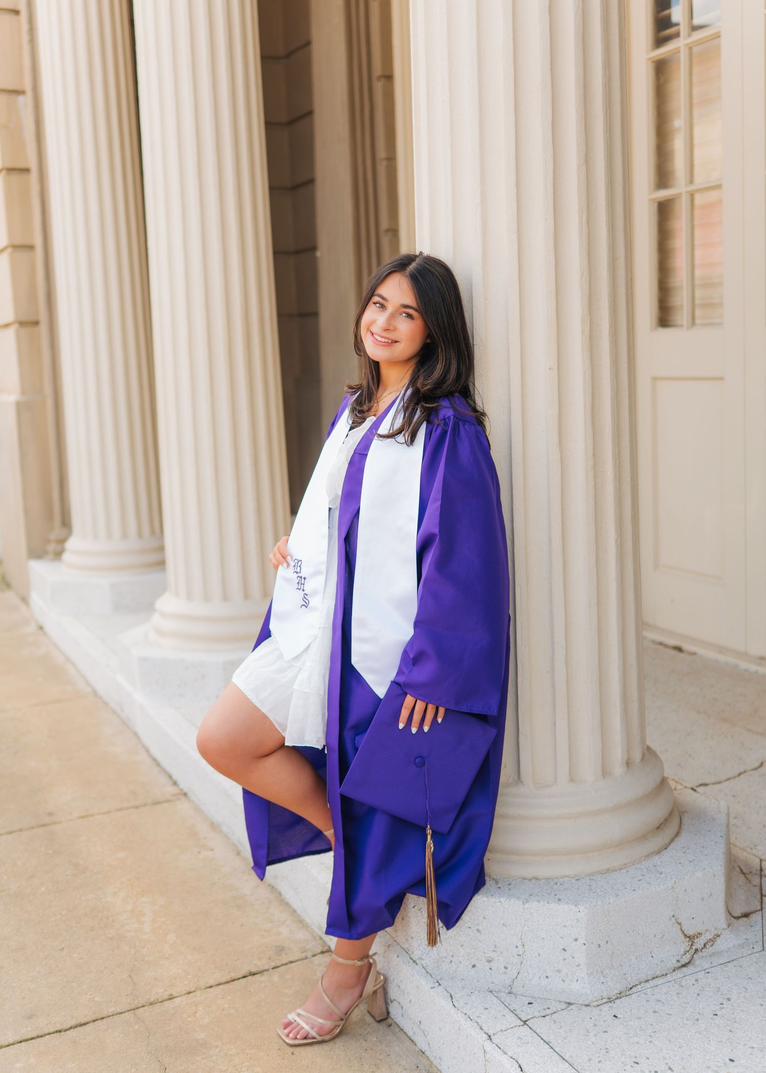 Graduate in purple cap and gown leaning against white column at university building entrance.