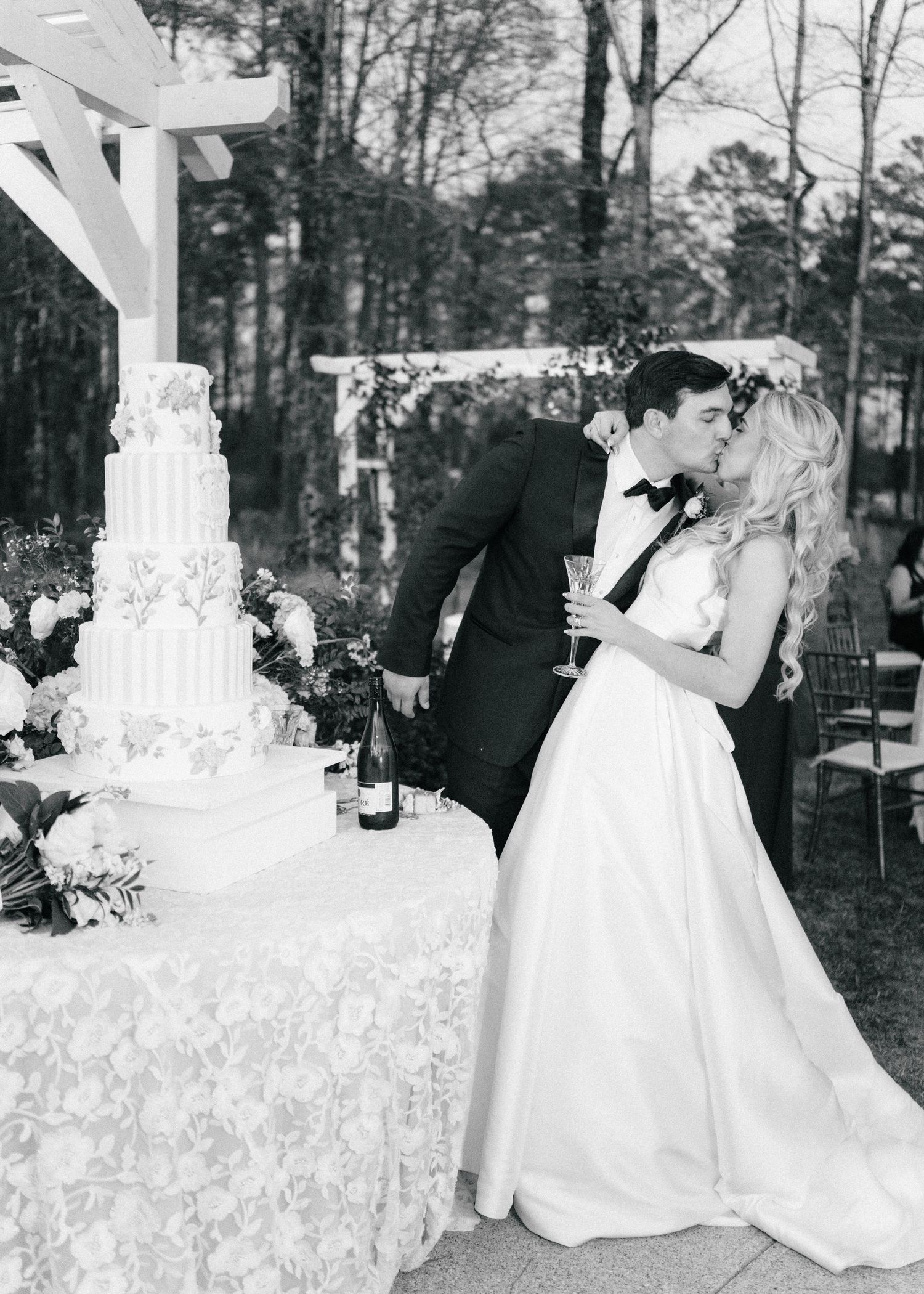 Black and white photo of bride and groom kissing beside elegant tiered wedding cake at outdoor reception.