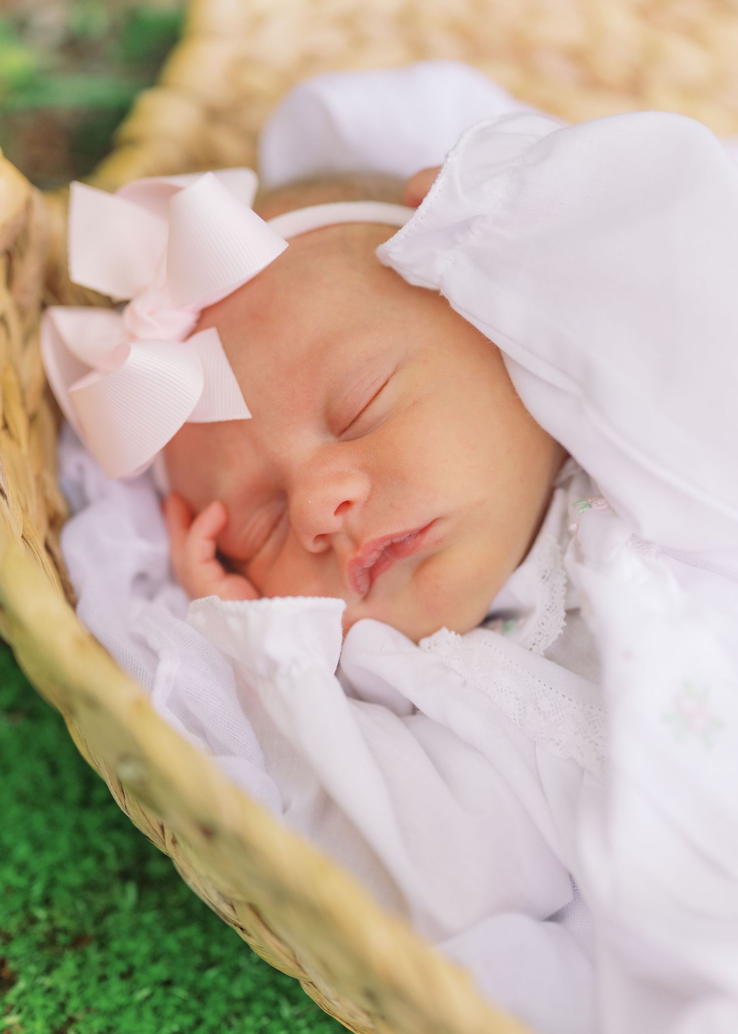 Sleeping newborn baby girl in white outfit with pink satin bow headband in wicker basket.