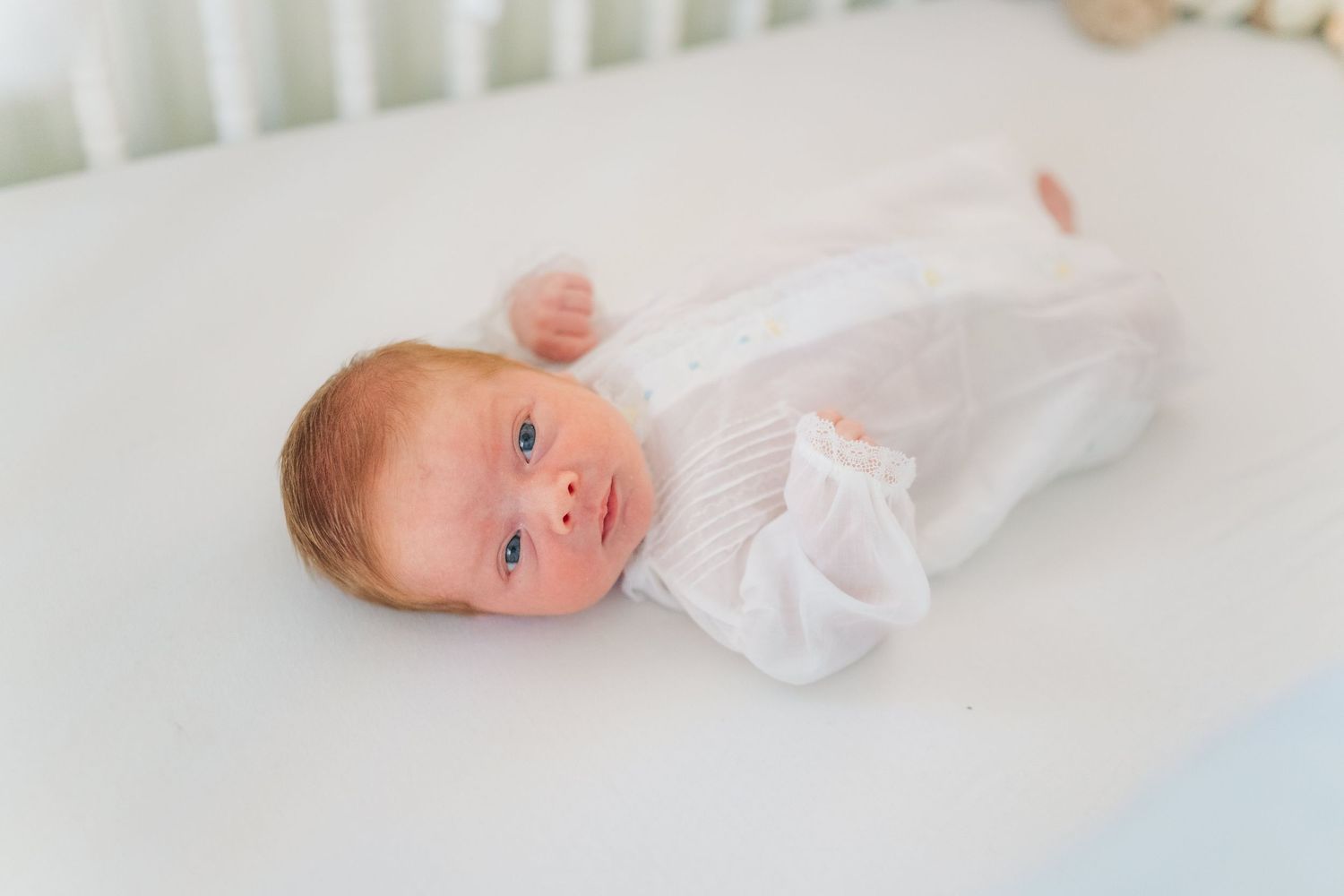 Newborn baby in white knit outfit lying in a white crib, looking up with blue eyes.