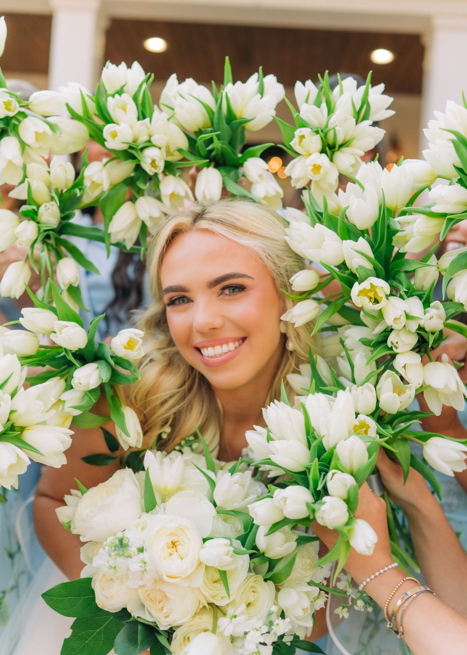 Smiling blonde bride framed by white tulip bouquets held by bridesmaids at outdoor wedding celebration.