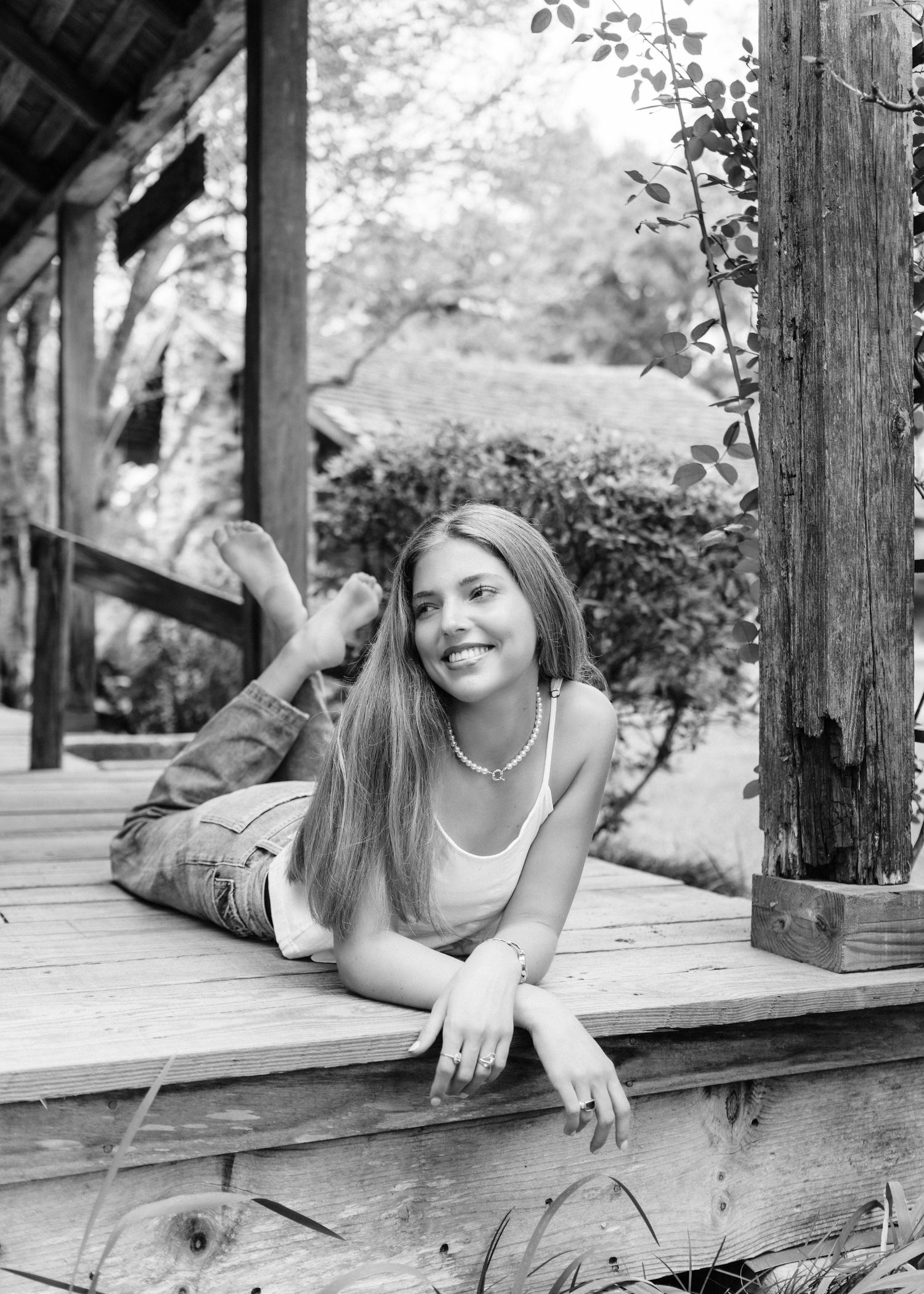 Smiling teen girl lies on wooden porch steps wearing jeans and tank top in black and white photo.