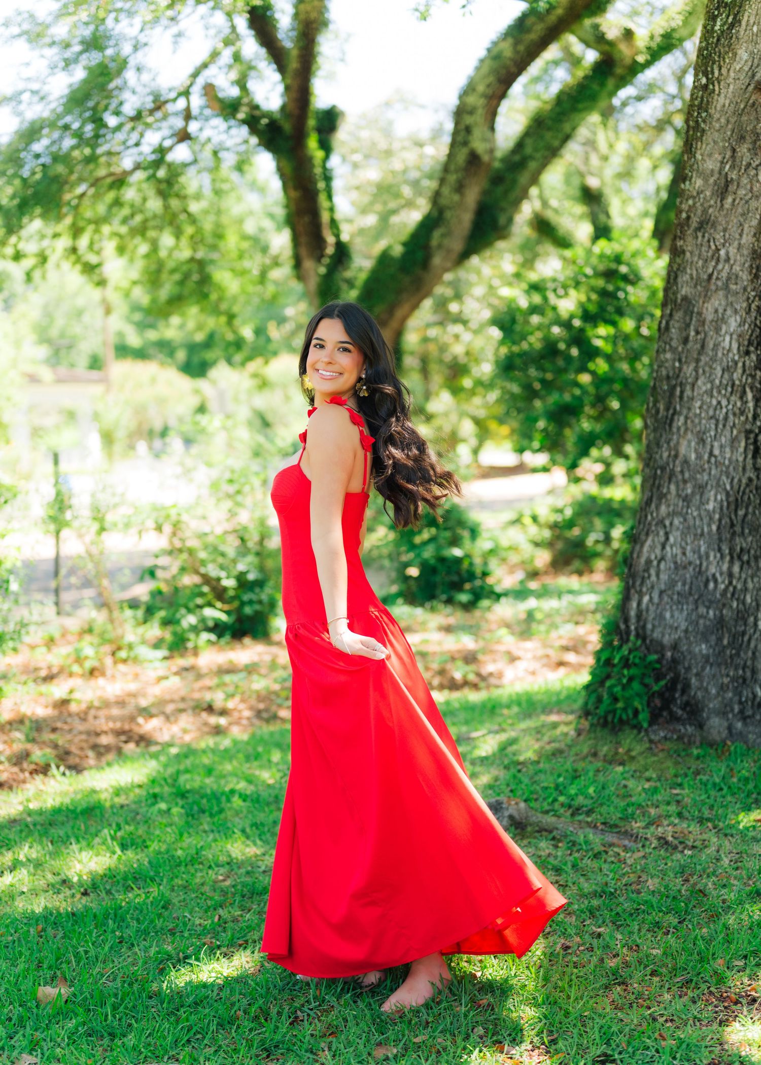 Woman in flowing red halter maxi dress twirls near large oak tree.