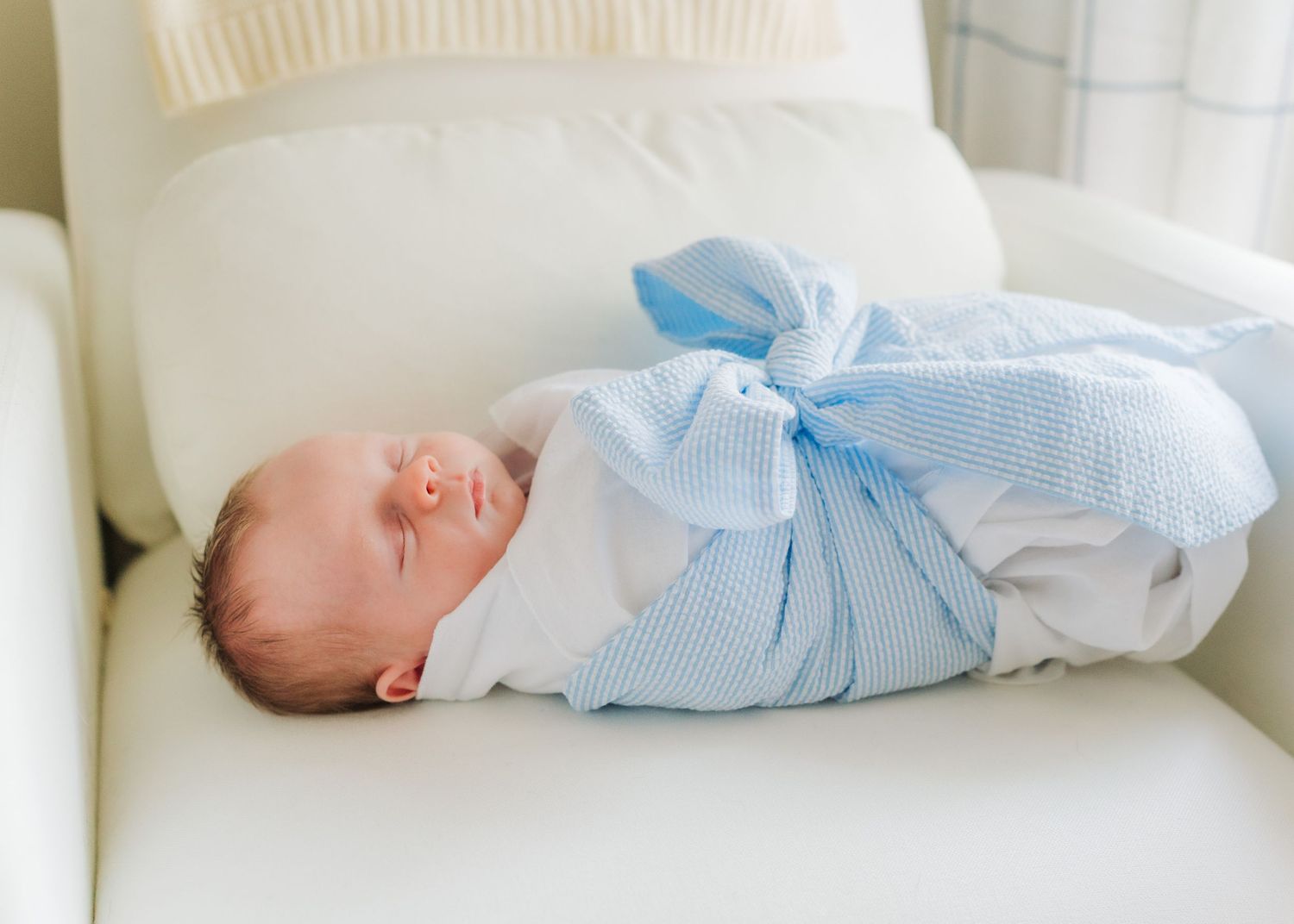 Newborn baby swaddled in a blue and white blanket sleeping peacefully on a cream white chair.