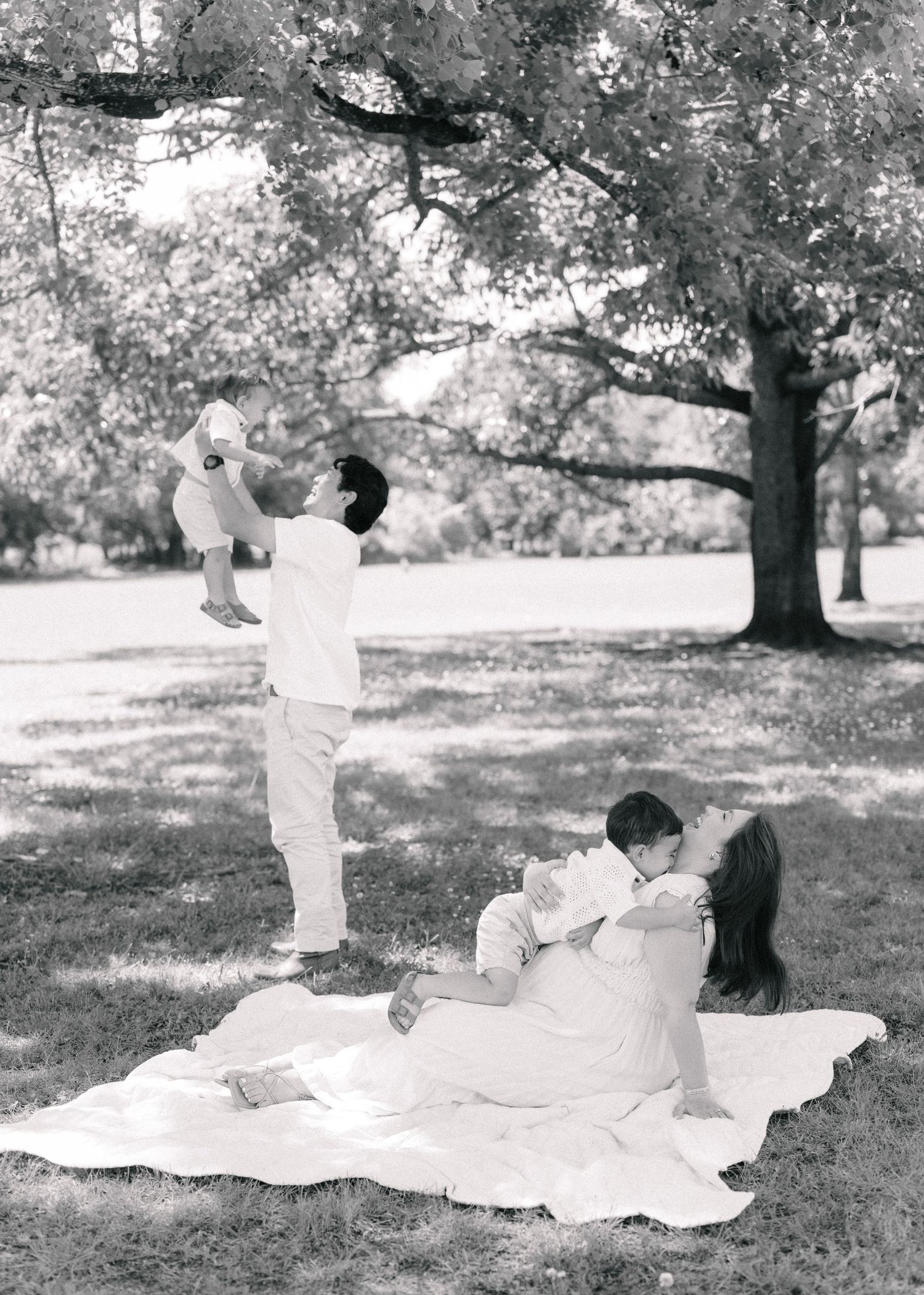 Black and white family photo on a blanket in the park, with a man lifting a baby while a woman holds another child.