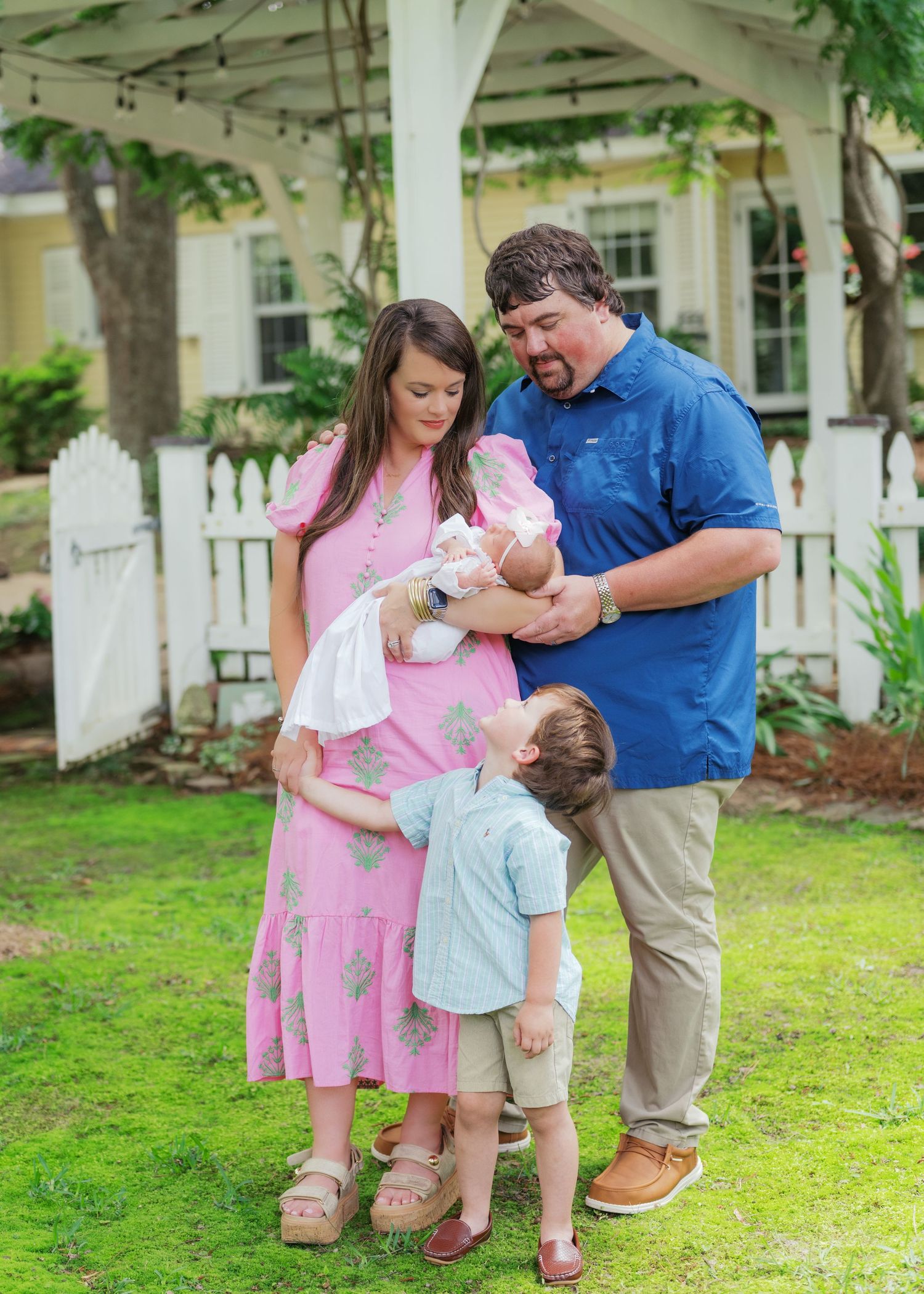 Family of four with newborn baby girl gathering under garden gazebo with white picket fence.
