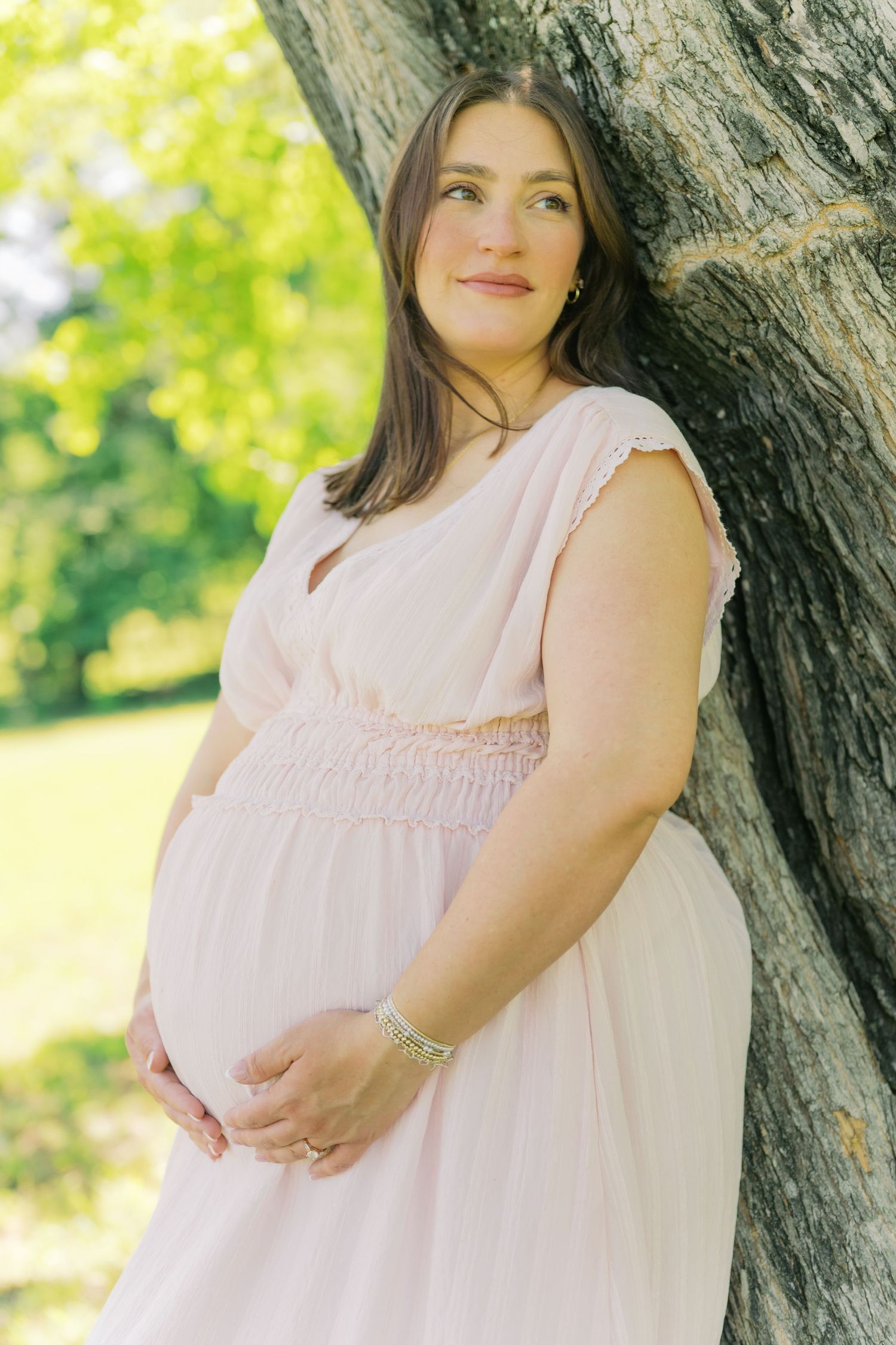 Pregnant woman in pink dress cradling baby bump while leaning against large tree in sunny outdoor park.