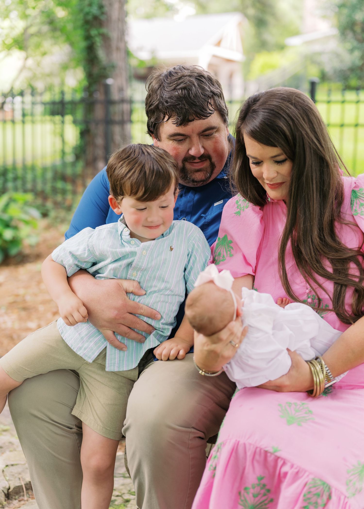 Parents and toddler son sitting outdoors admiring tiny newborn baby girl's feet in pink dress.