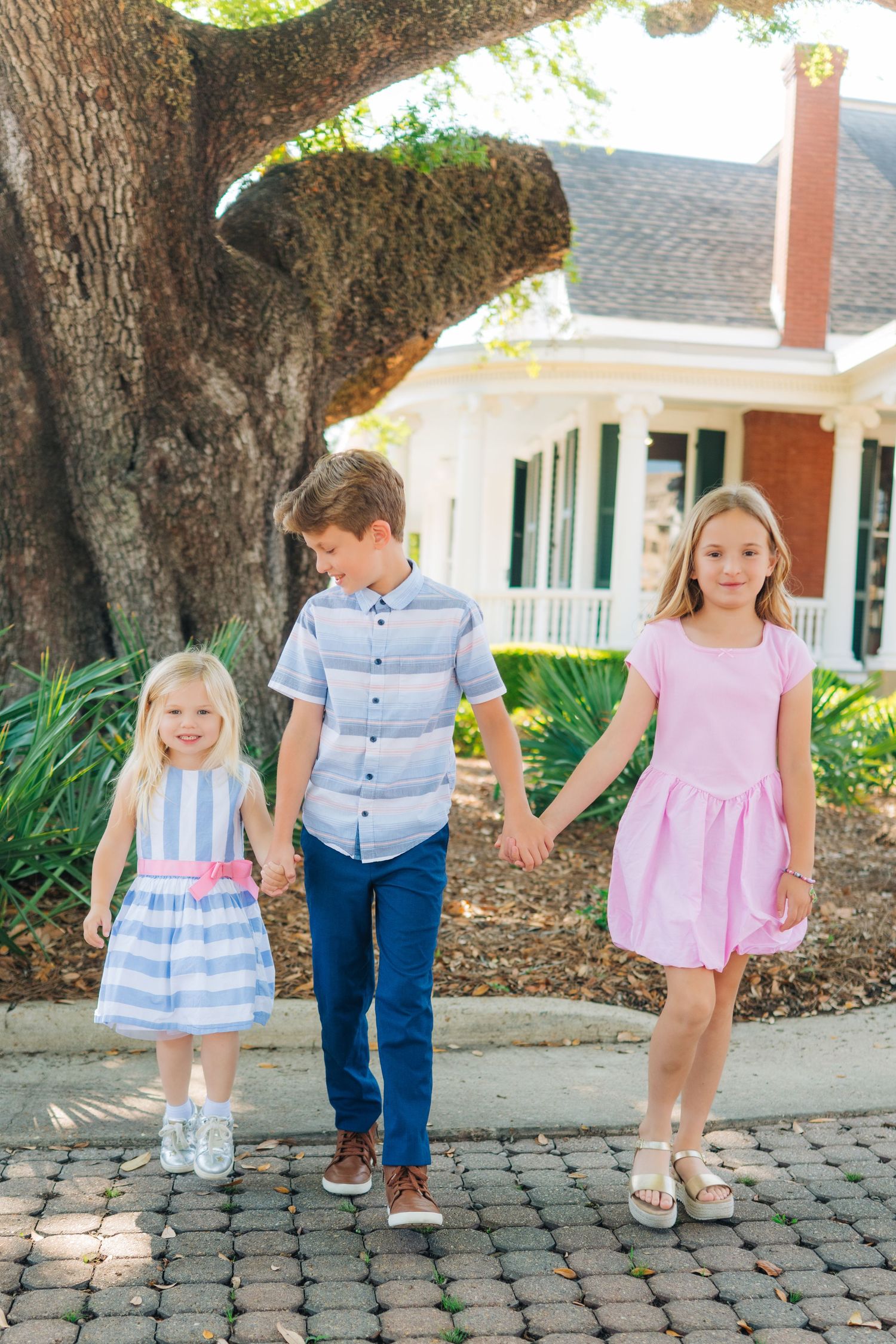 Three children holding hands outdoors, two girls in pink and blue dresses and a boy in a blue shirt and navy pants.