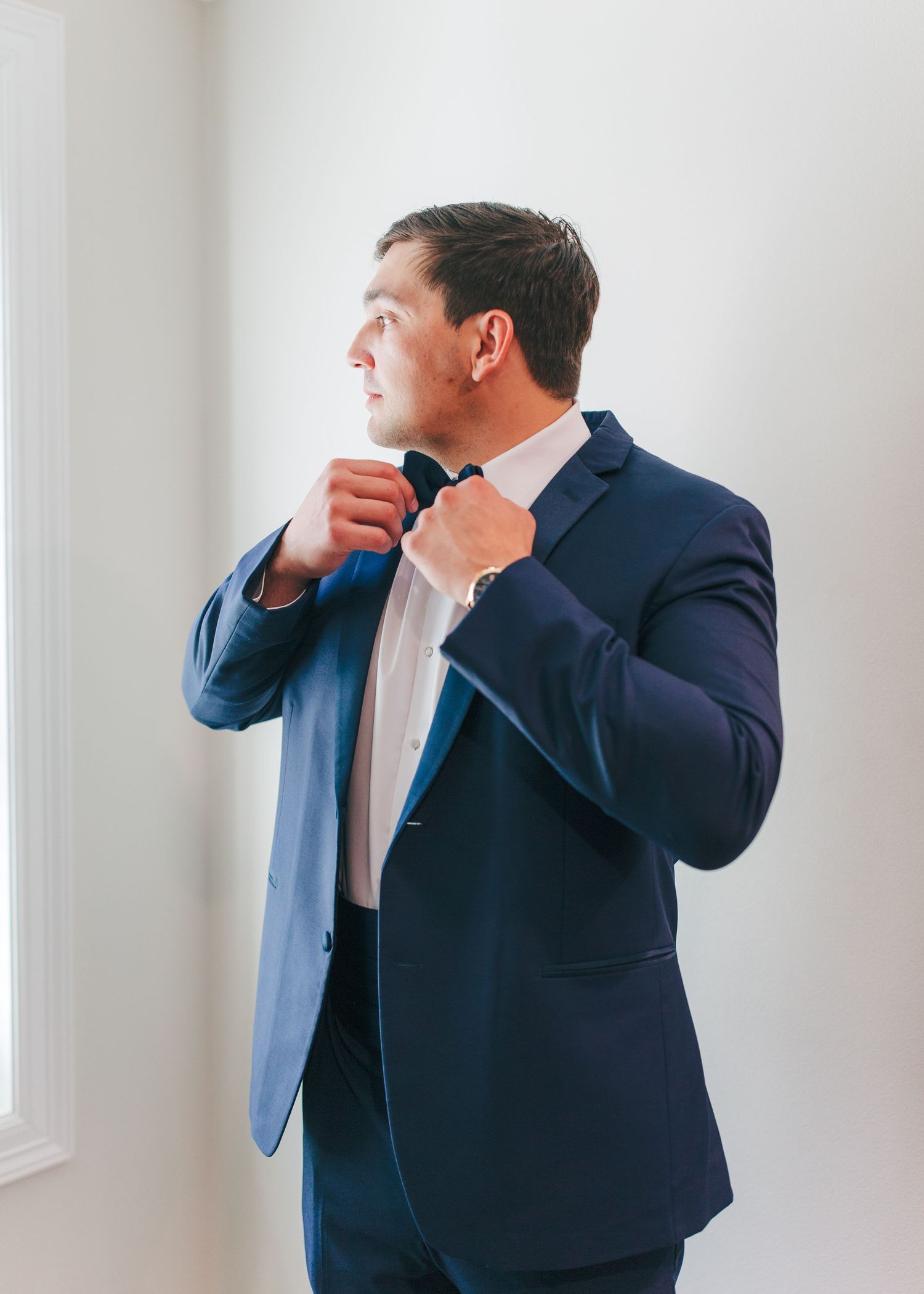 Groom in navy suit adjusting bow tie while looking out window on wedding day.