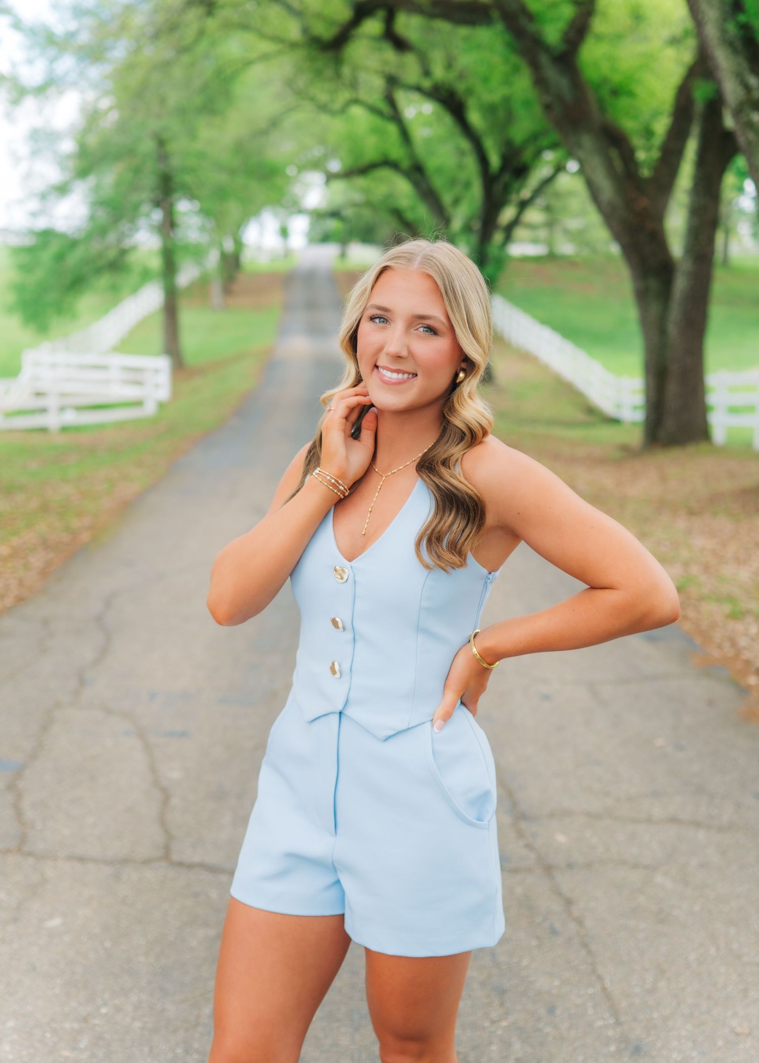 Young blonde woman in light blue romper poses on a tree-lined pathway near a white fence on a sunny day.