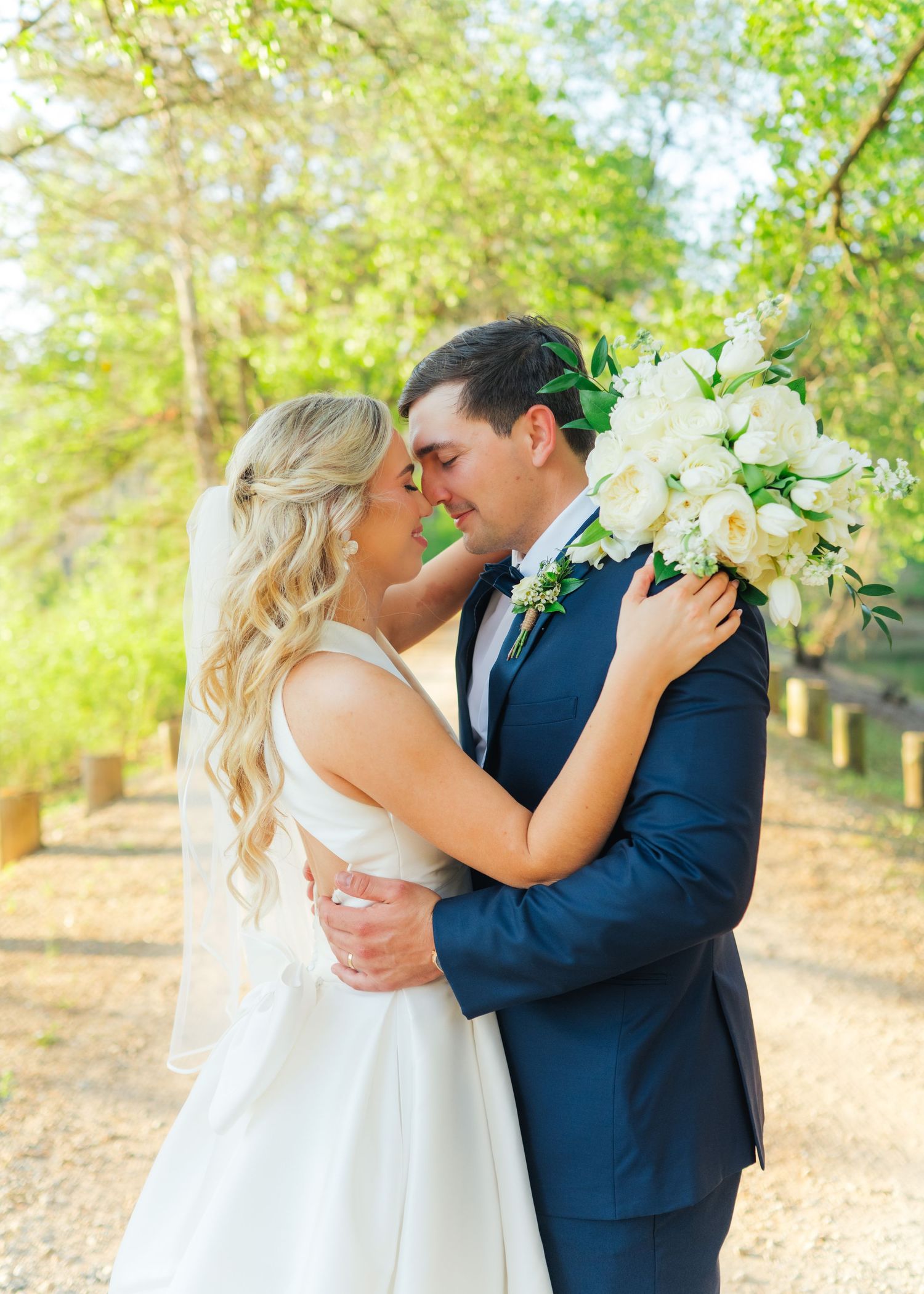 Bride and groom share romantic moment outdoors, she holds white floral bouquet as they lean in for a kiss.