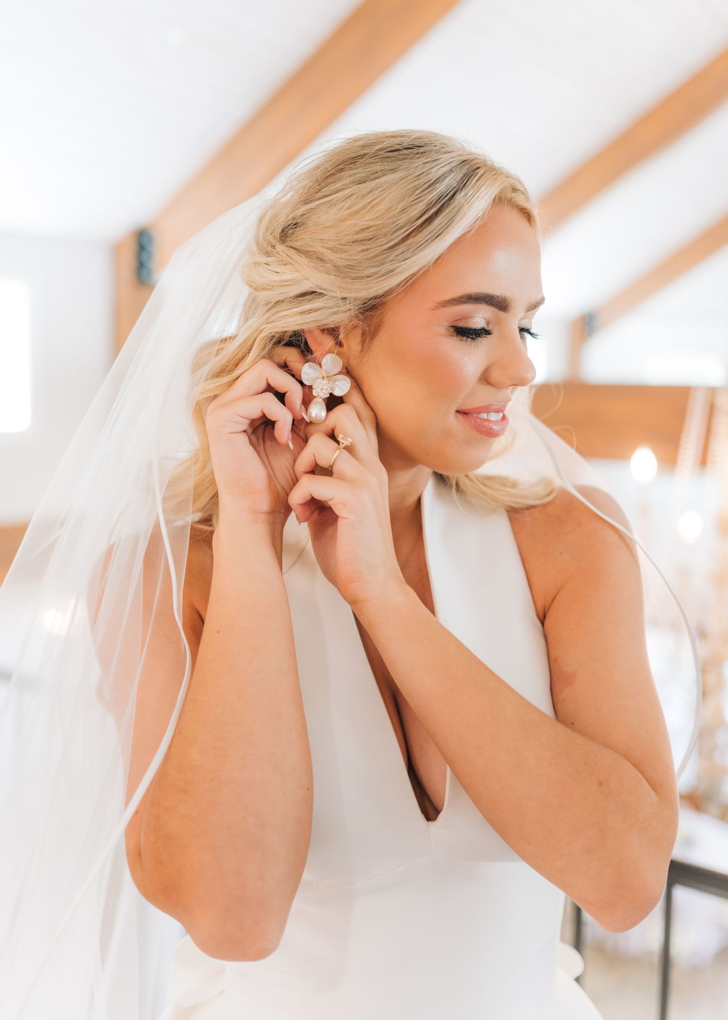 Blonde bride in white dress and veil putting on earrings while getting ready on her wedding day.