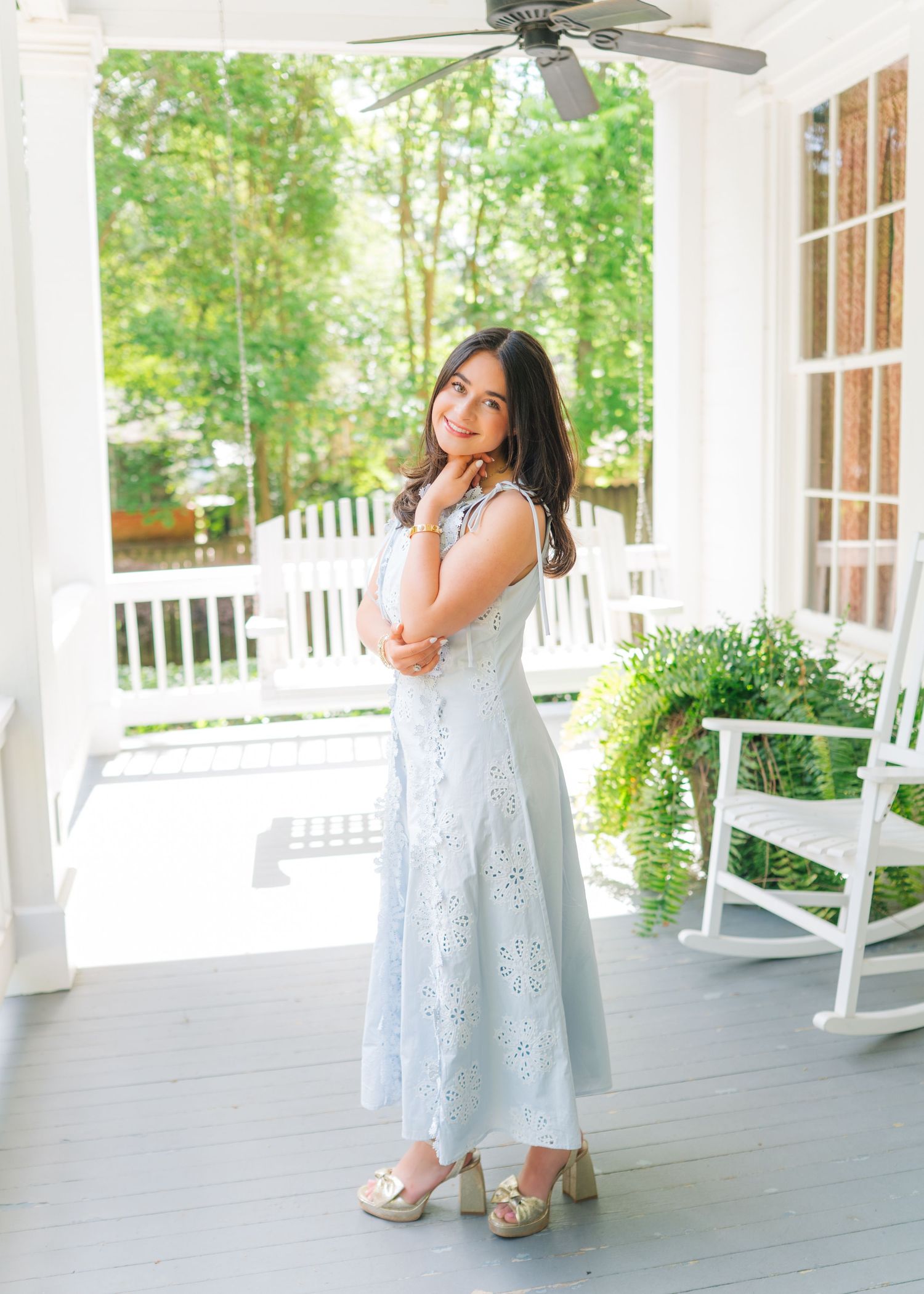 Young woman in light blue eyelet midi dress and gold heels posing on a white Southern porch.