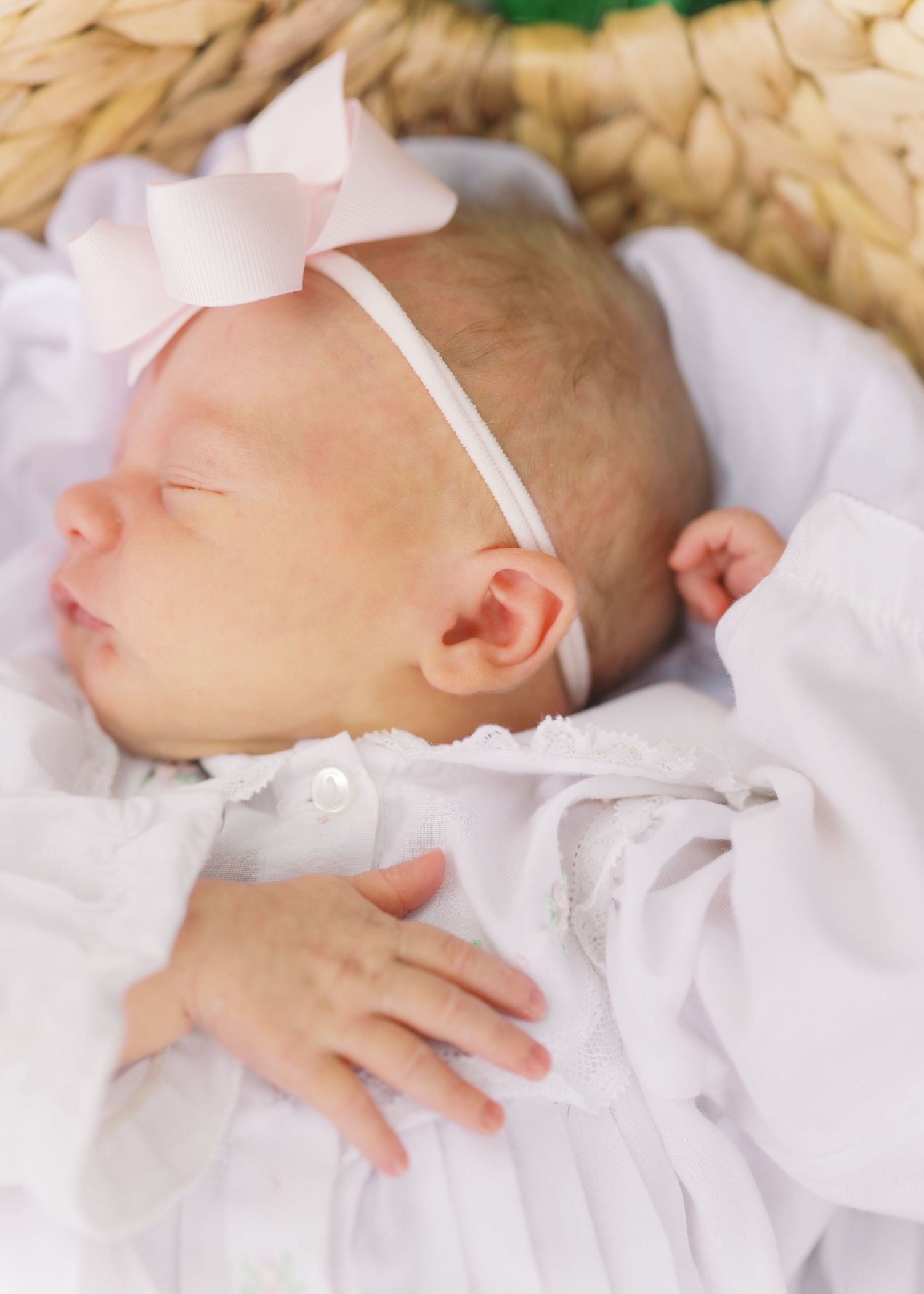 Sleeping newborn baby wearing white outfit and pink bow headband resting in wicker basket.