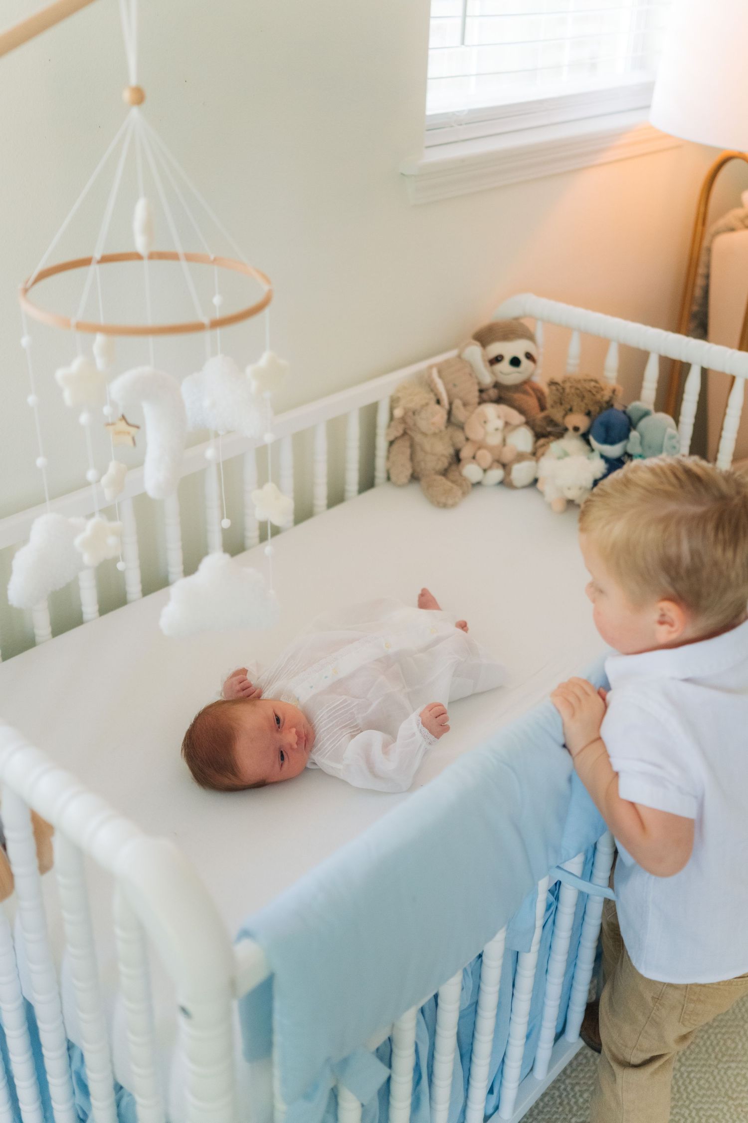 A toddler boy in a white shirt looks over a white crib at his newborn sibling surrounded by stuffed animals.